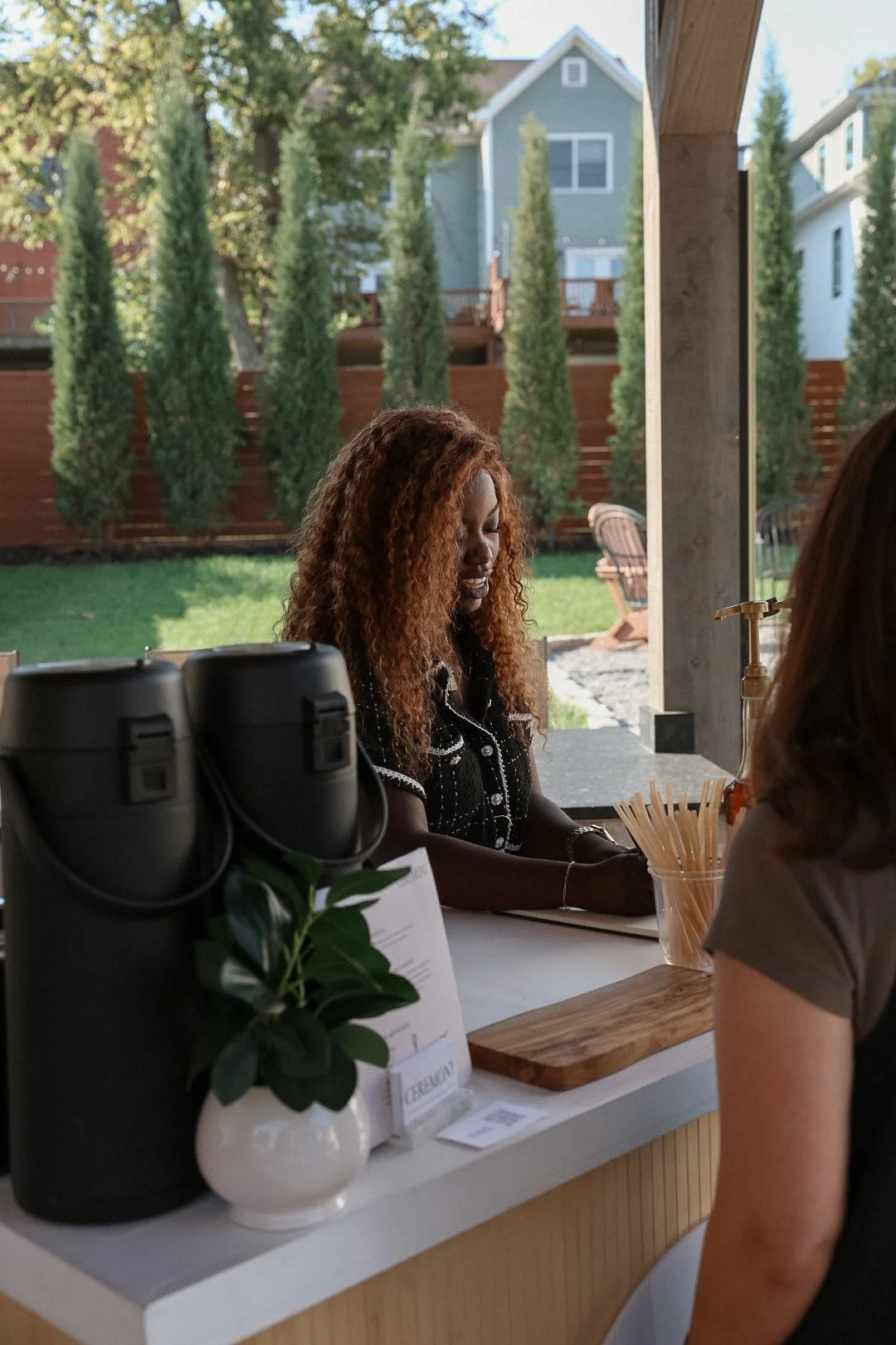 A woman with curly hair working at a counter at a matcha cart, with large windows showing an outdoor green yard with trees and houses in the background.