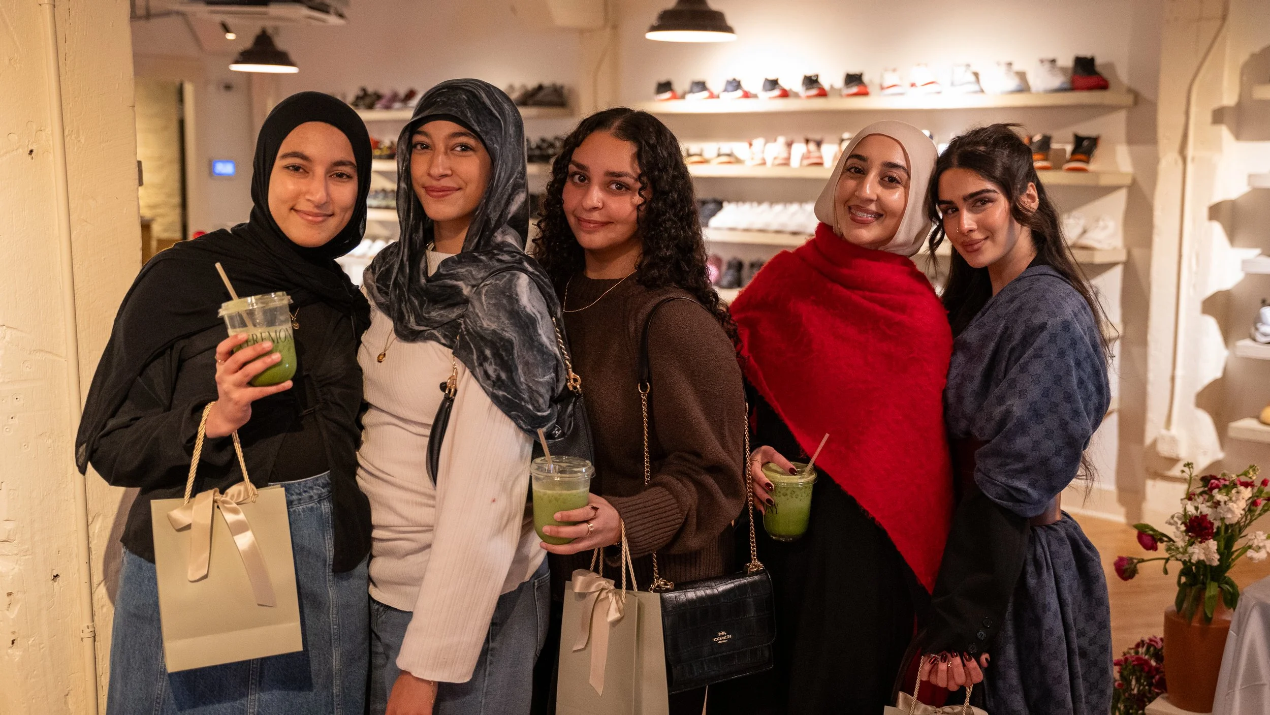 Five women standing in a store, holding green drinks, smiling at the camera, with shelves of shoes behind them.