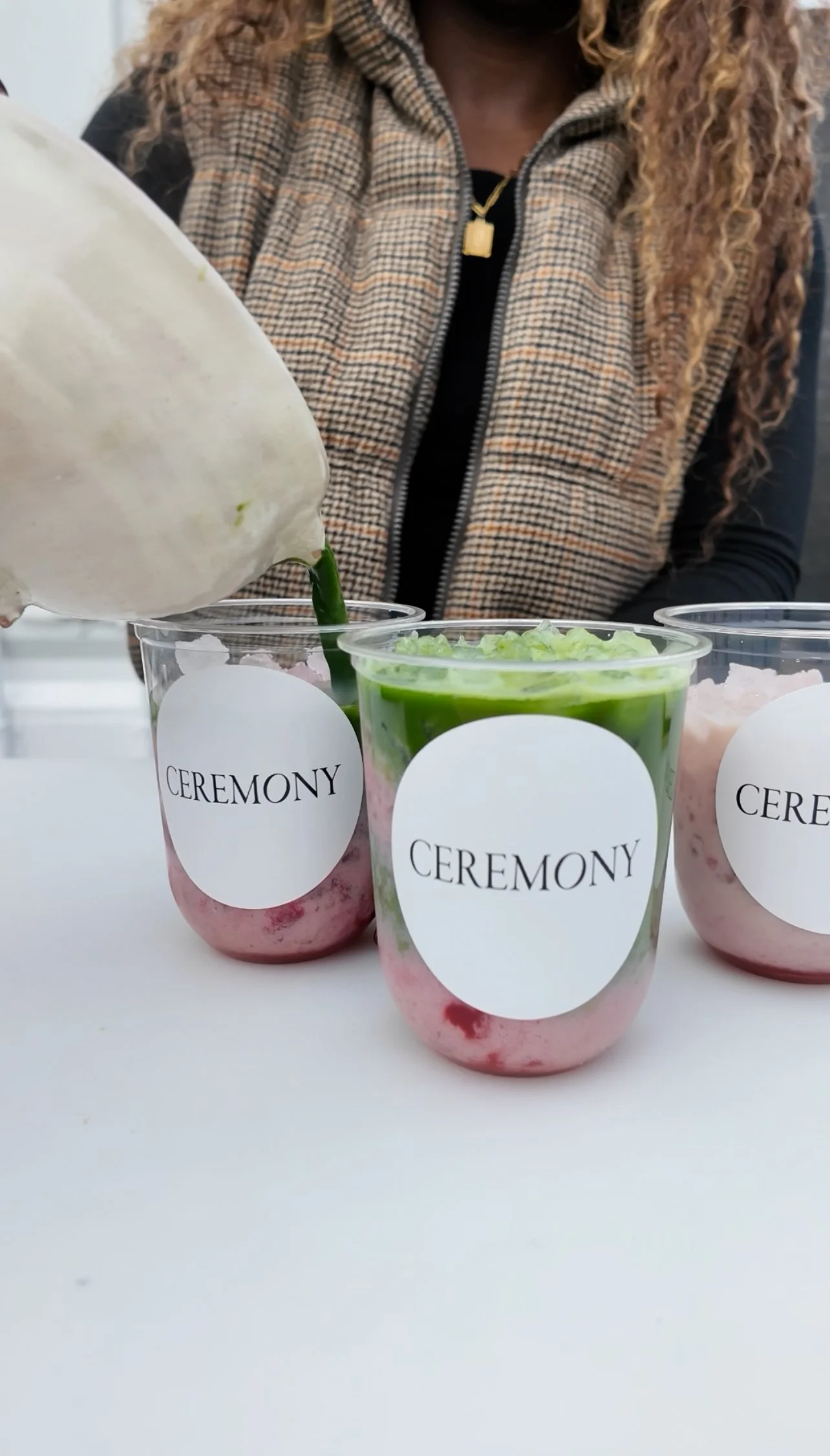 Dessert cups labeled 'CEREMONY' with pink and green layered desserts and a woman in a plaid vest pouring a white substance into one of the cups.