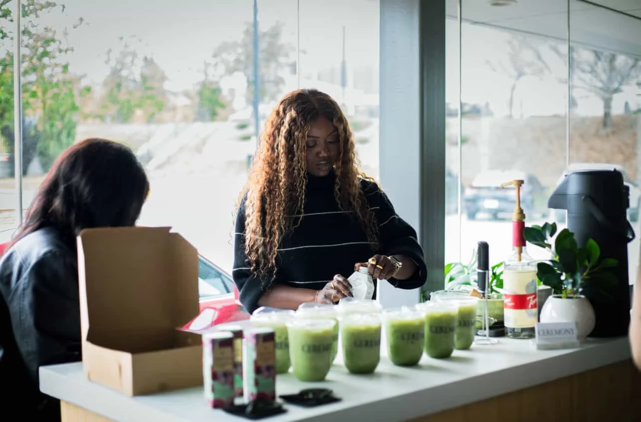 A woman with curly hair preparing green smoothies behind a counter in a bright cafe with large windows.