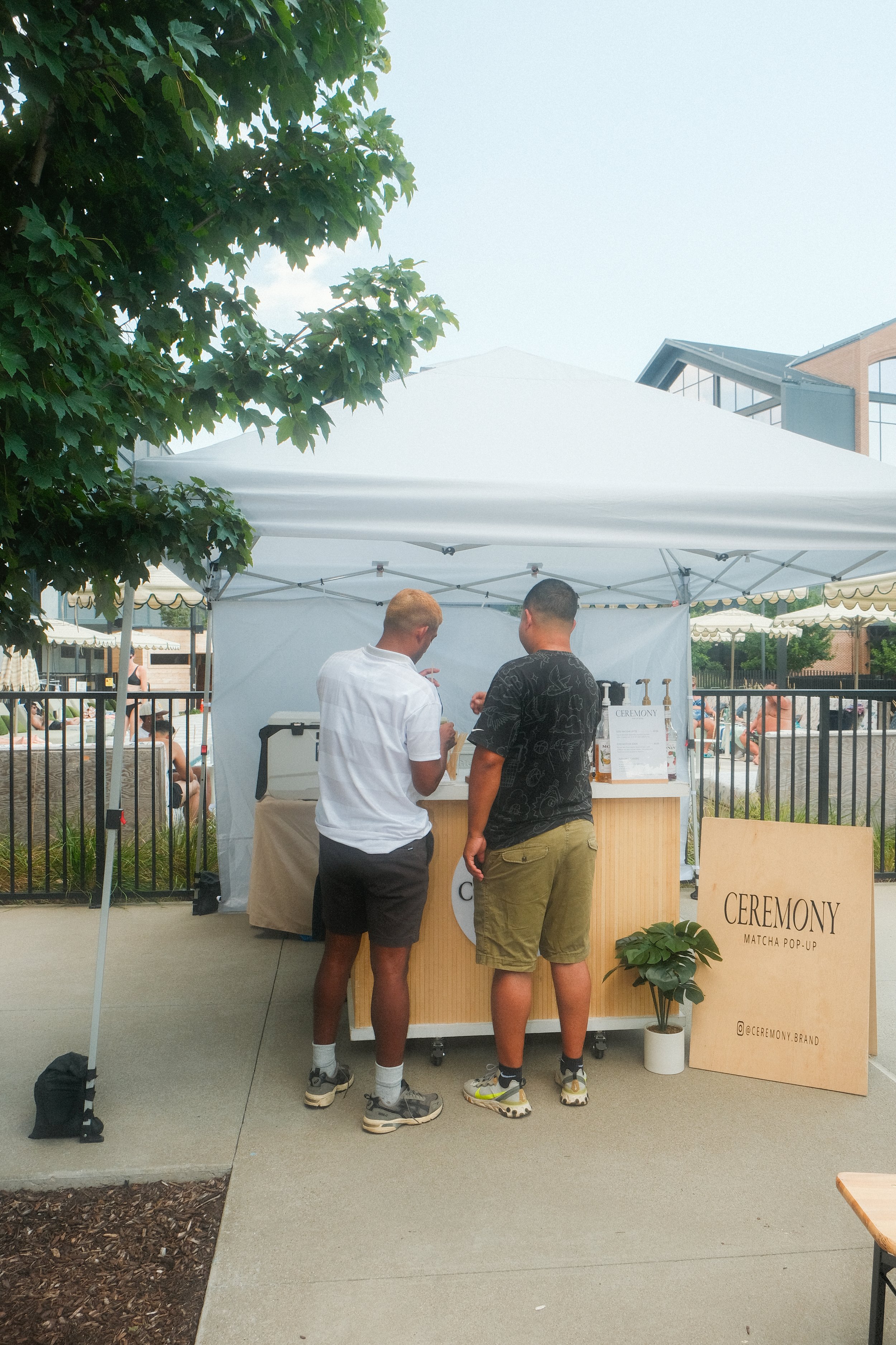 Two men standing at a small pop-up booth with a canopy, labeled 'CEREMONY,' serving matcha drinks outdoors at a public event or park.
