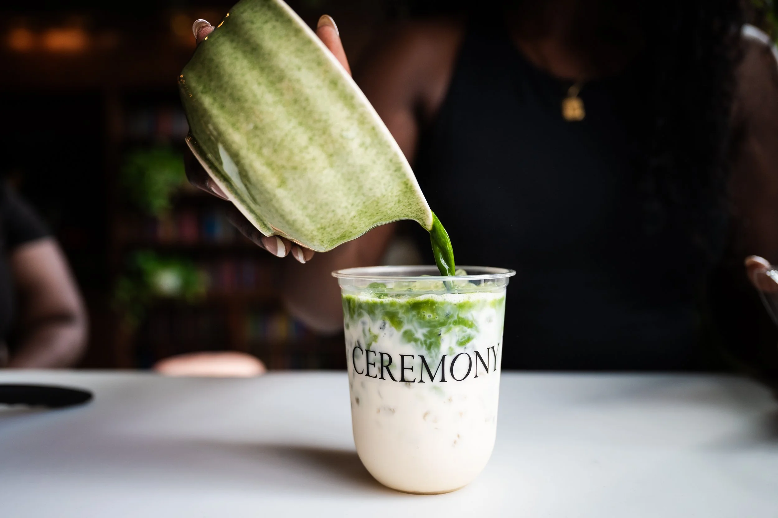 A person pours a green matcha latte from a green bowl into a plastic cup labeled 'CEREMONY', with ice visible inside the drink.