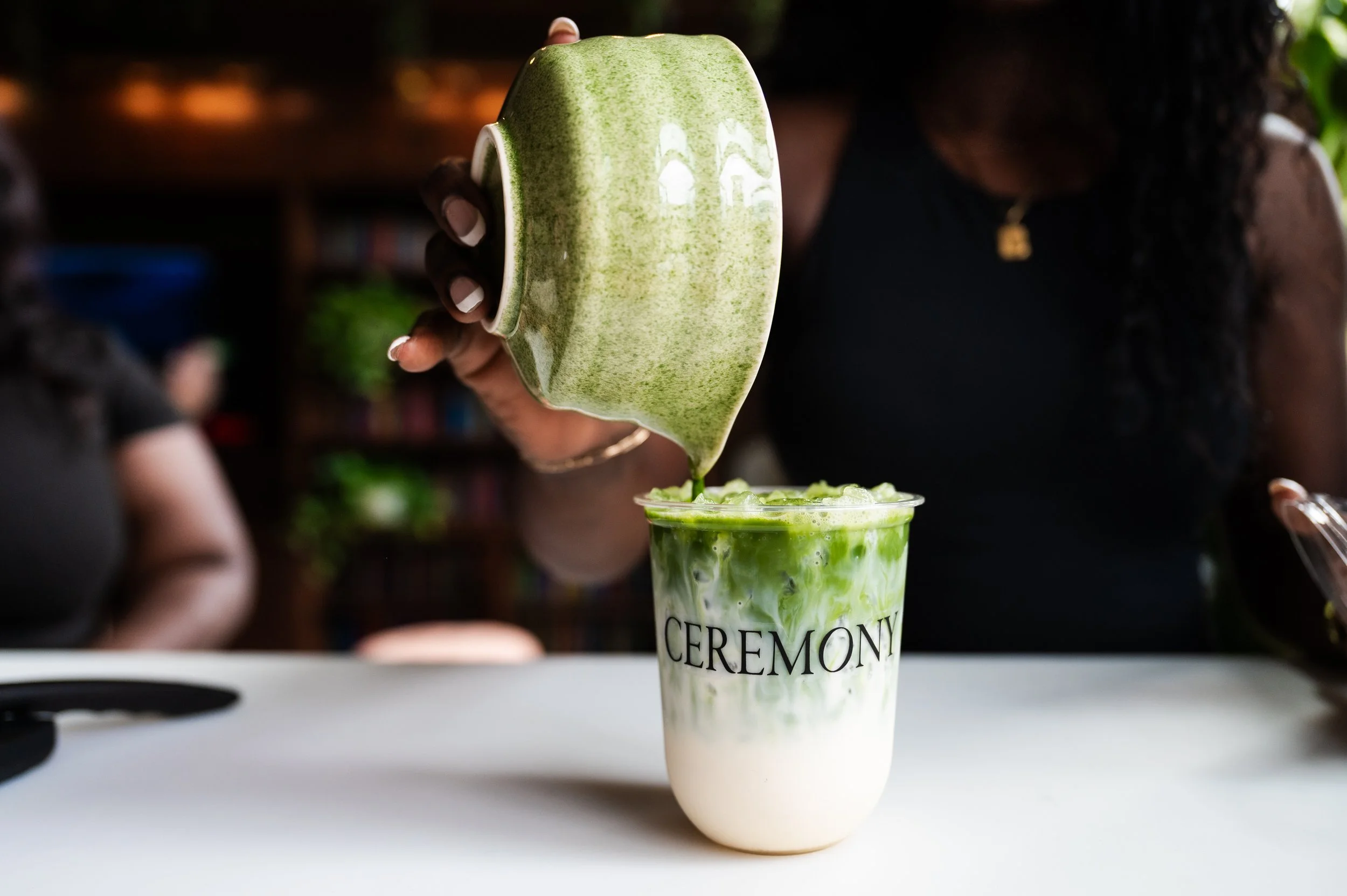 Person pouring a green matcha latte with milk into a glass with the word 'CEREMONY' printed on it.