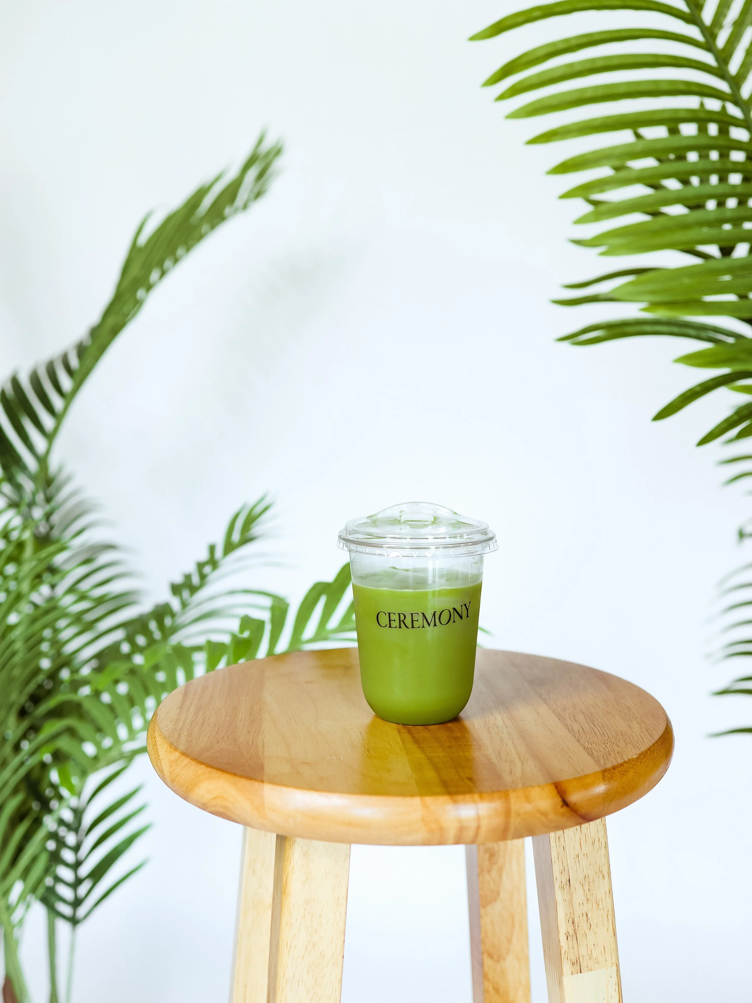 A green beverage in a clear plastic cup with a lid, labeled 'CEREMONY,' placed on a round wooden stool, flanked by green foliage.