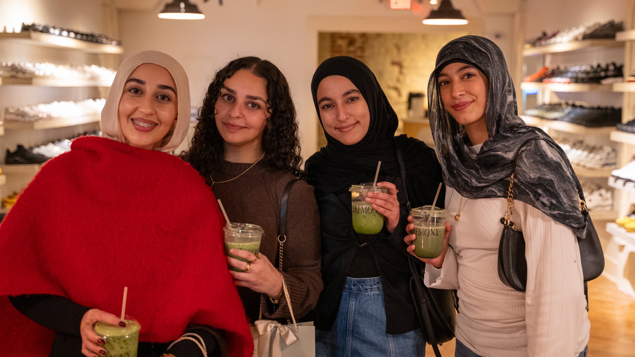 Four young women standing in a shoe store, smiling, and holding green drinks with straws. The store has shelves with various shoes in the background.
