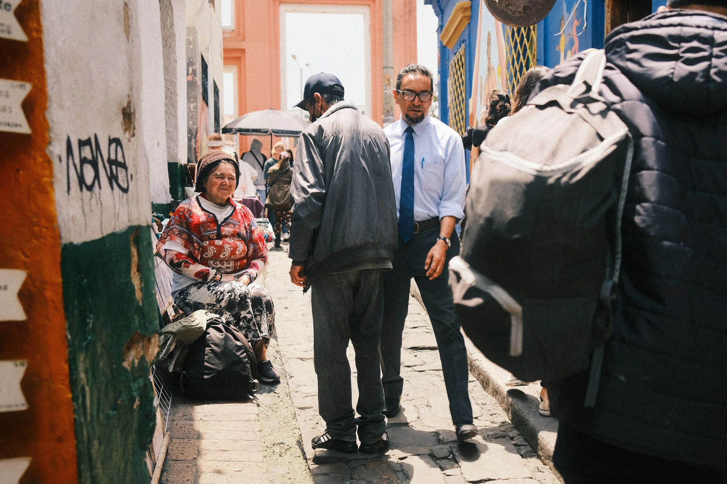 Street scene with multiple people walking and talking in a colorful area with murals, a woman sitting on the curb, and a man with a backpack in the foreground.