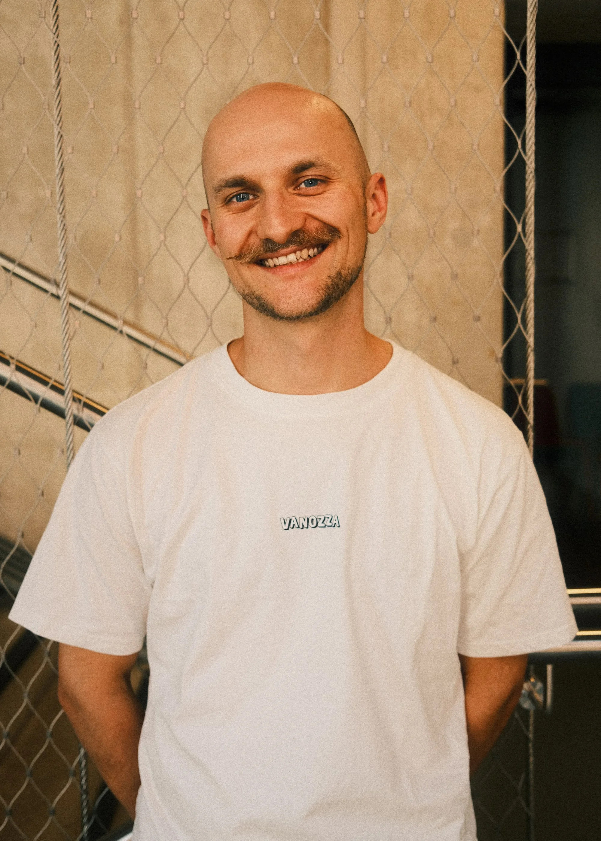 A smiling man with a bald head, a mustache, and beard, wearing a white t-shirt with "VANOZZA" written on it, standing in front of a chain-link fence.