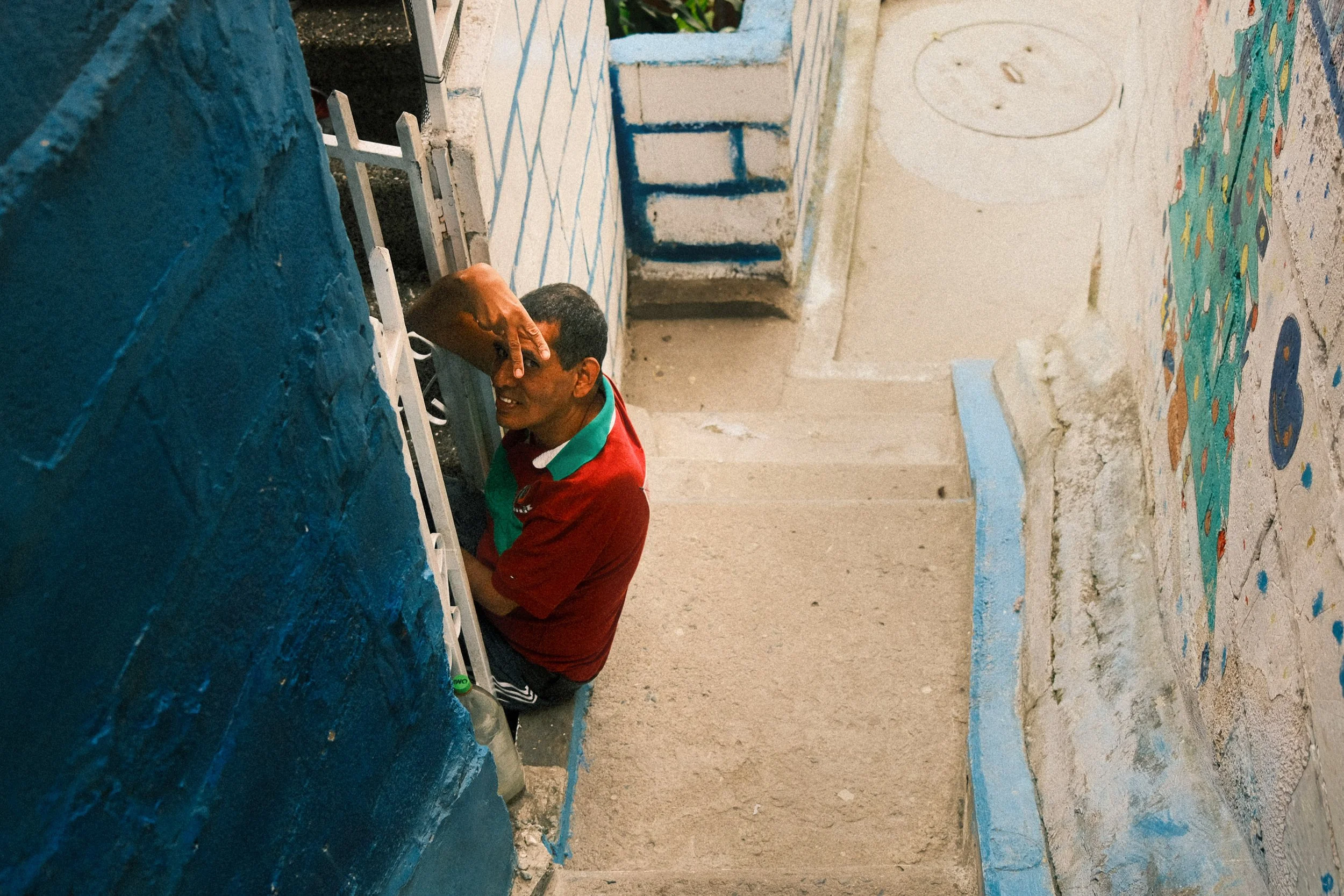 A man in a red shirt sits on stairs next to a blue and white painted wall, looking up with one hand shielding his eyes from the light.