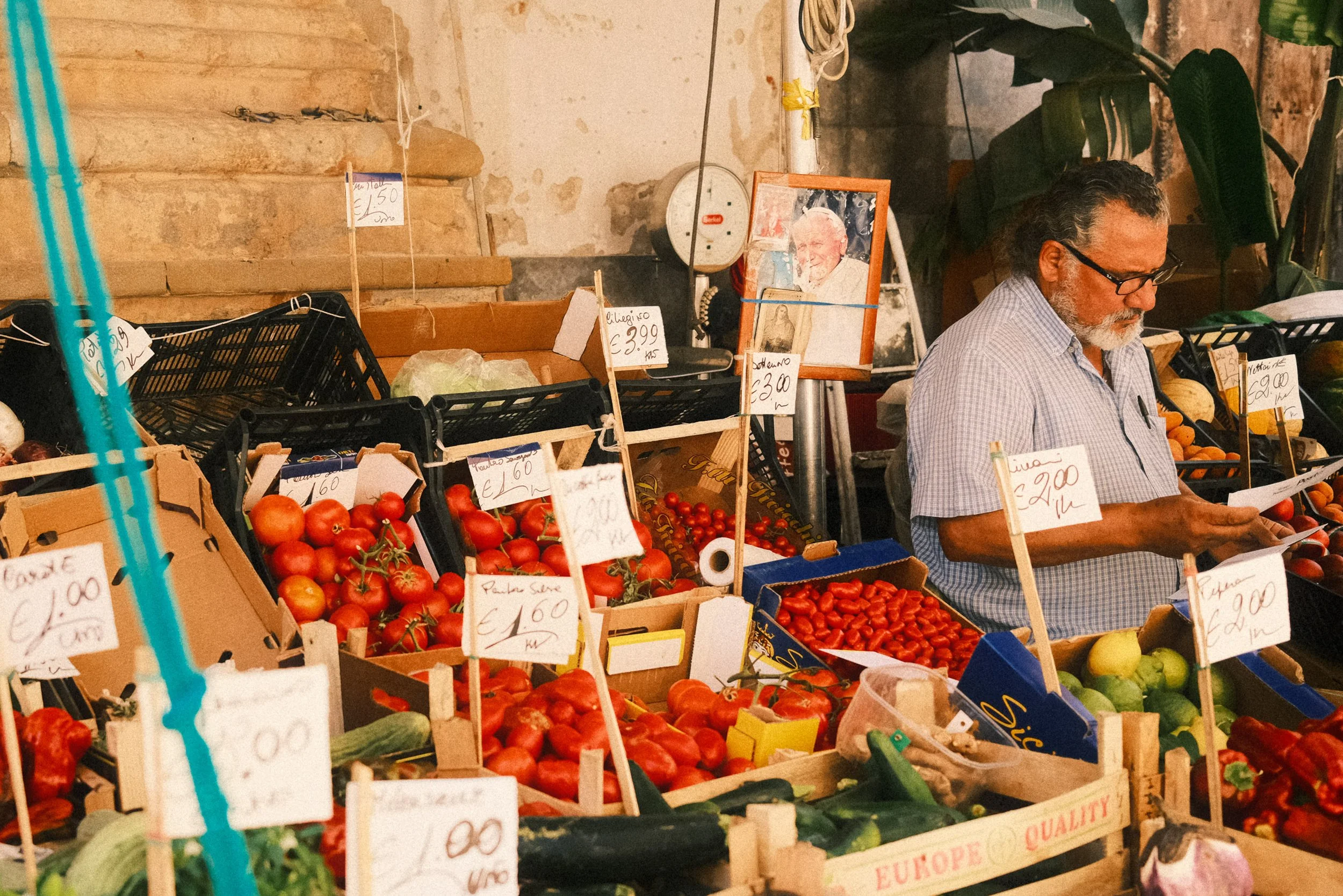 A man wearing glasses and a striped shirt shopping at a fruit and vegetable stall in a marketplace. The stall has tomatoes, cucumbers, zucchinis, and other produce with price tags, and a framed photo of a man in the background.