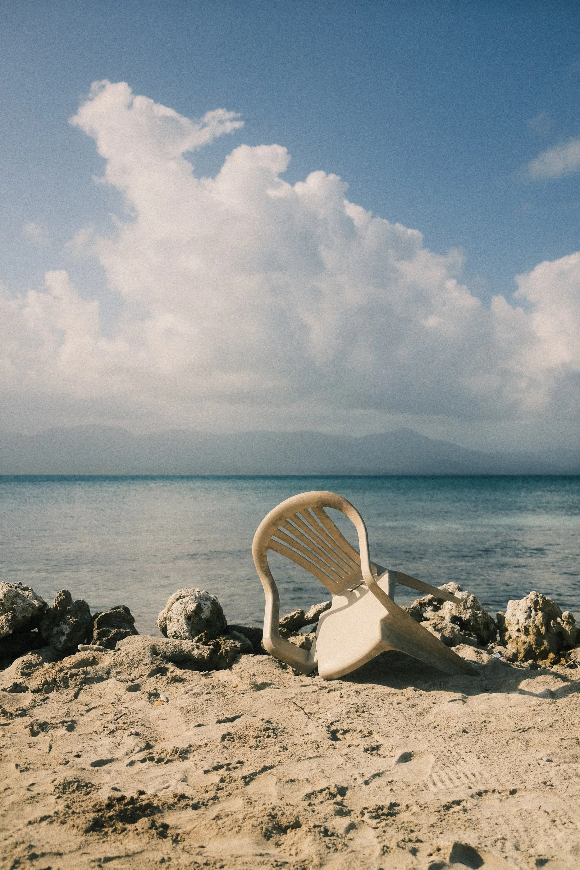 A white plastic chair tipped over on sandy beach with rocks, overlooking calm ocean waters and cloudy sky in the background.