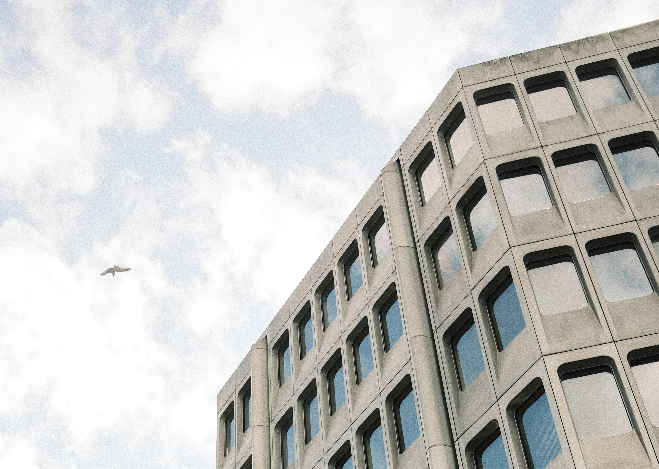 A modern building with a geometric facade and multiple windows, against a partly cloudy sky with a bird flying nearby.