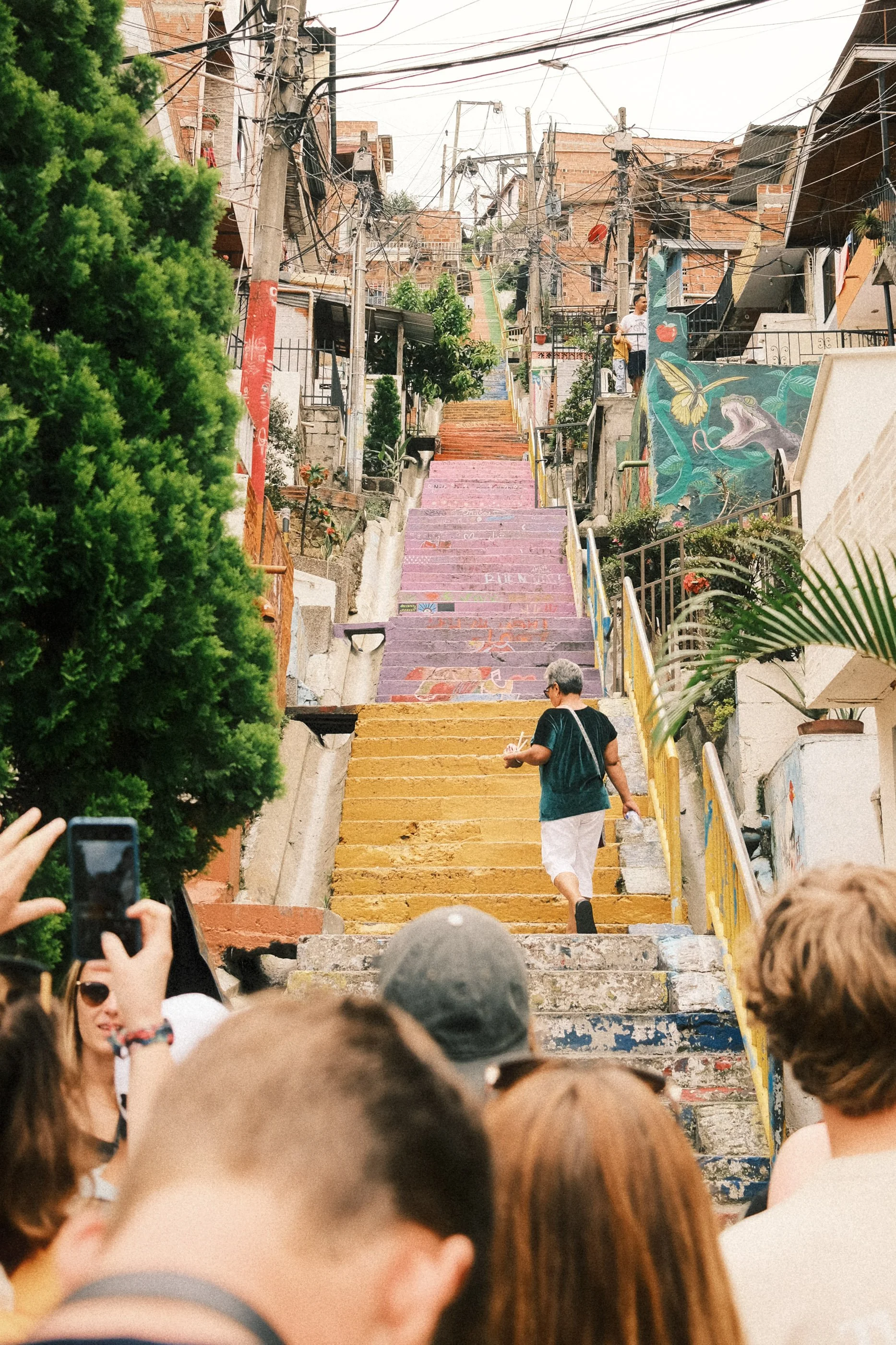 Colorful staircase in a neighborhood with murals, graffiti, and vegetation, with people taking photos.