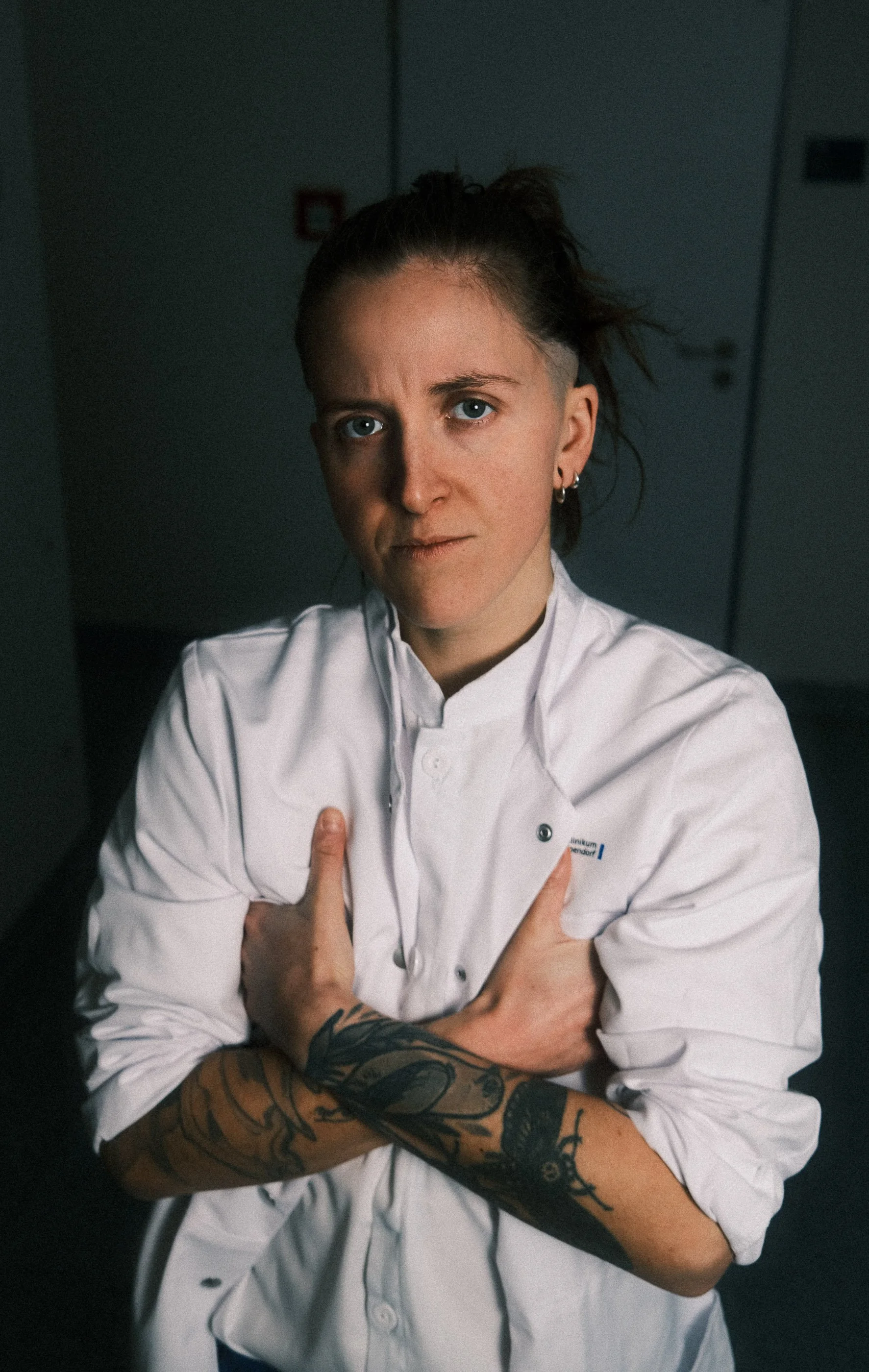 A person with tattoos on their arms, wearing a white hospital coat, standing indoors with her arms crossed and looking directly at the camera.