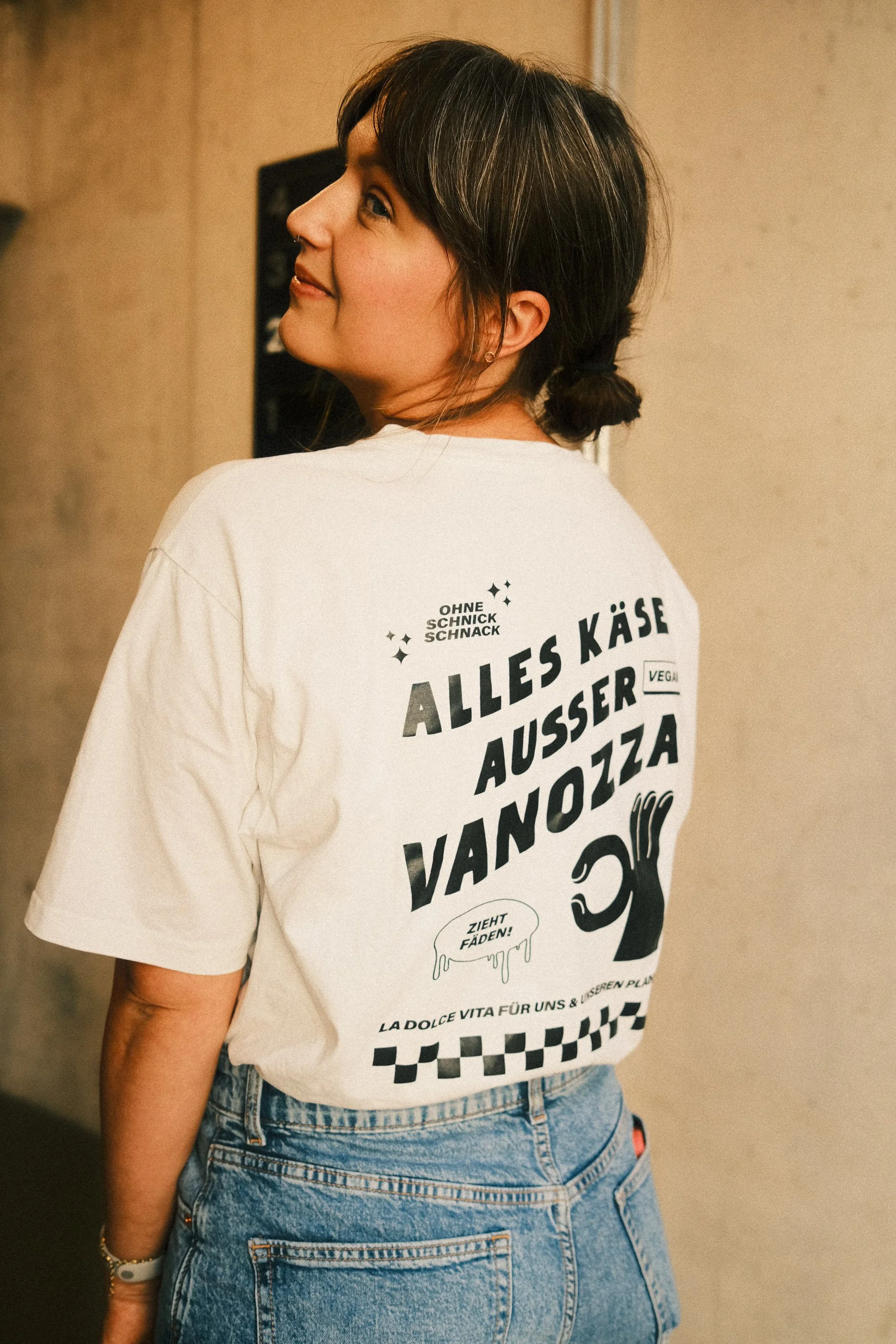A woman with dark hair tied in a bun, wearing a white graphic T-shirt and blue jeans, is standing with her back facing the camera, looking over her shoulder.