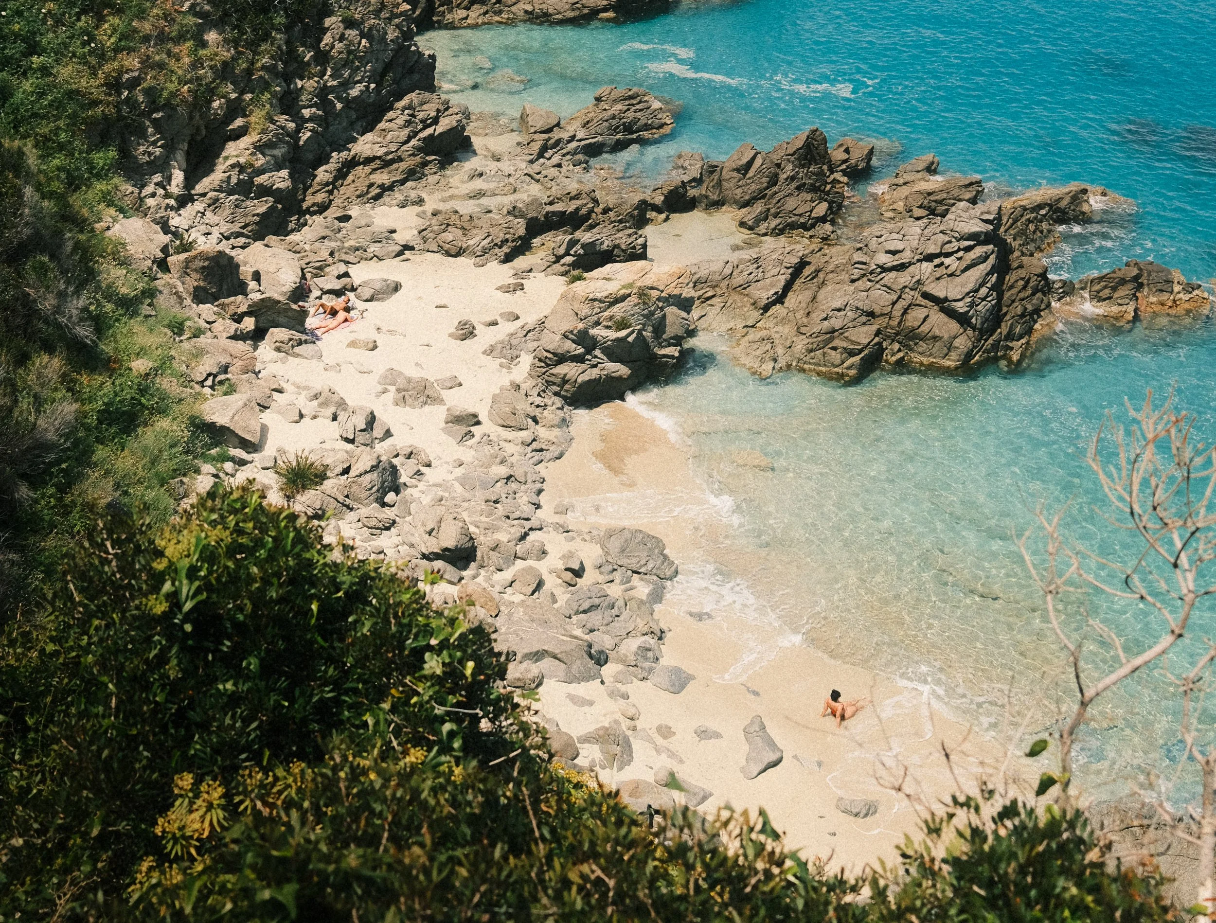 View of a secluded beach with light-colored sand, large rocks, and turquoise water, with greenery in the foreground and a few people relaxing on the beach.