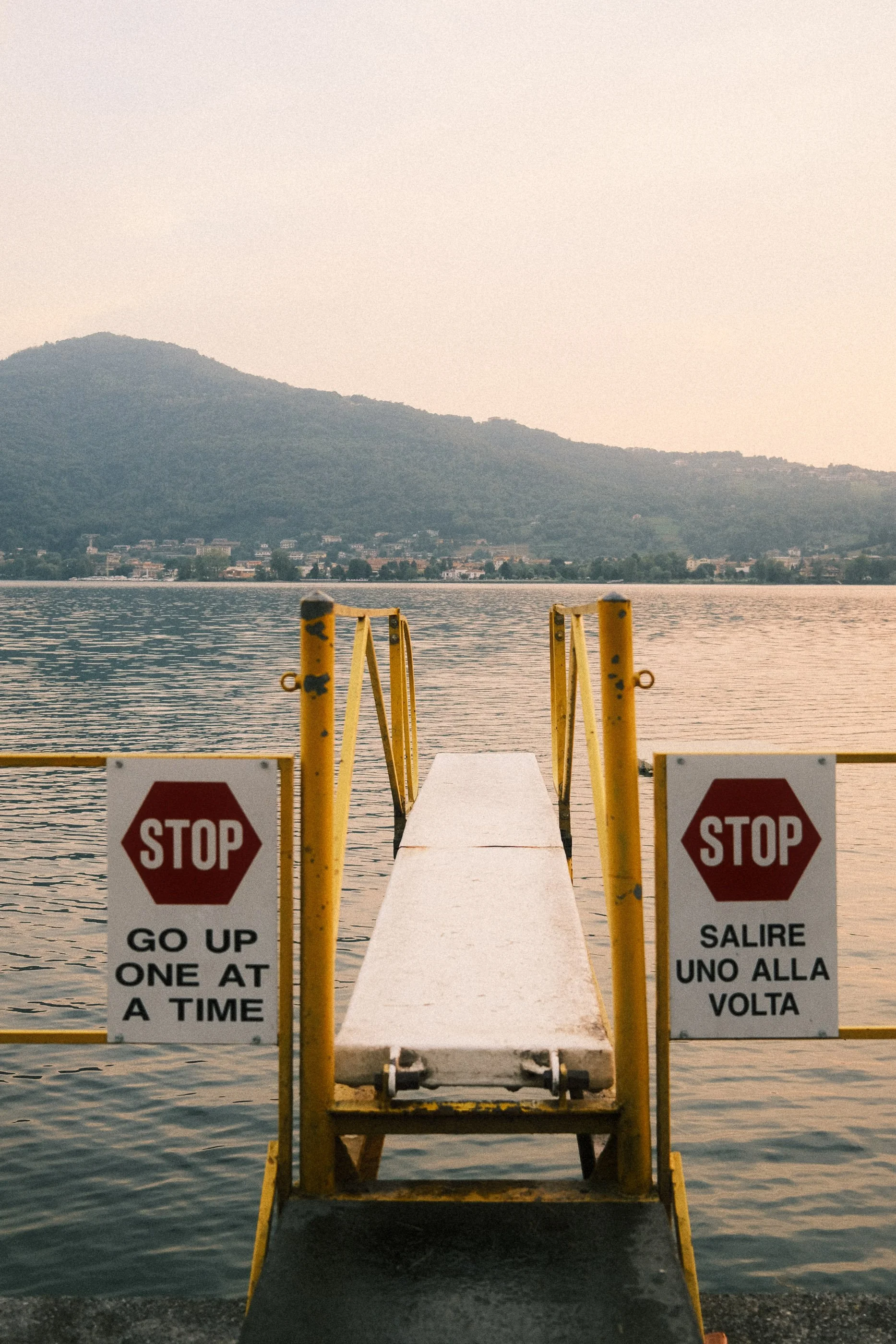 A small pier with yellow railings extending into a body of water, with two stop signs warning to go up one at a time. Mountains in the background and a calm lake.