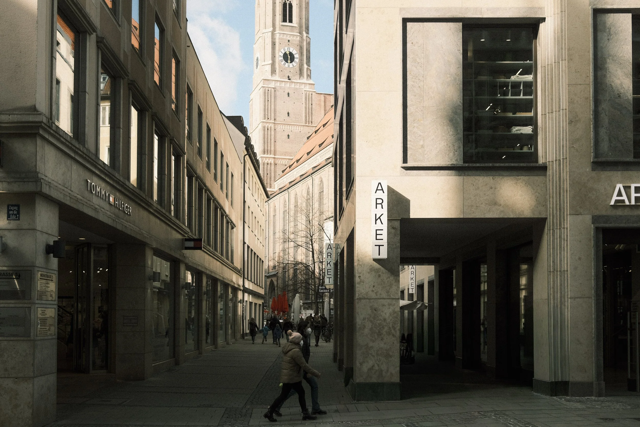 City street view showing modern and historic buildings, people walking, with a tall clock tower in the background.