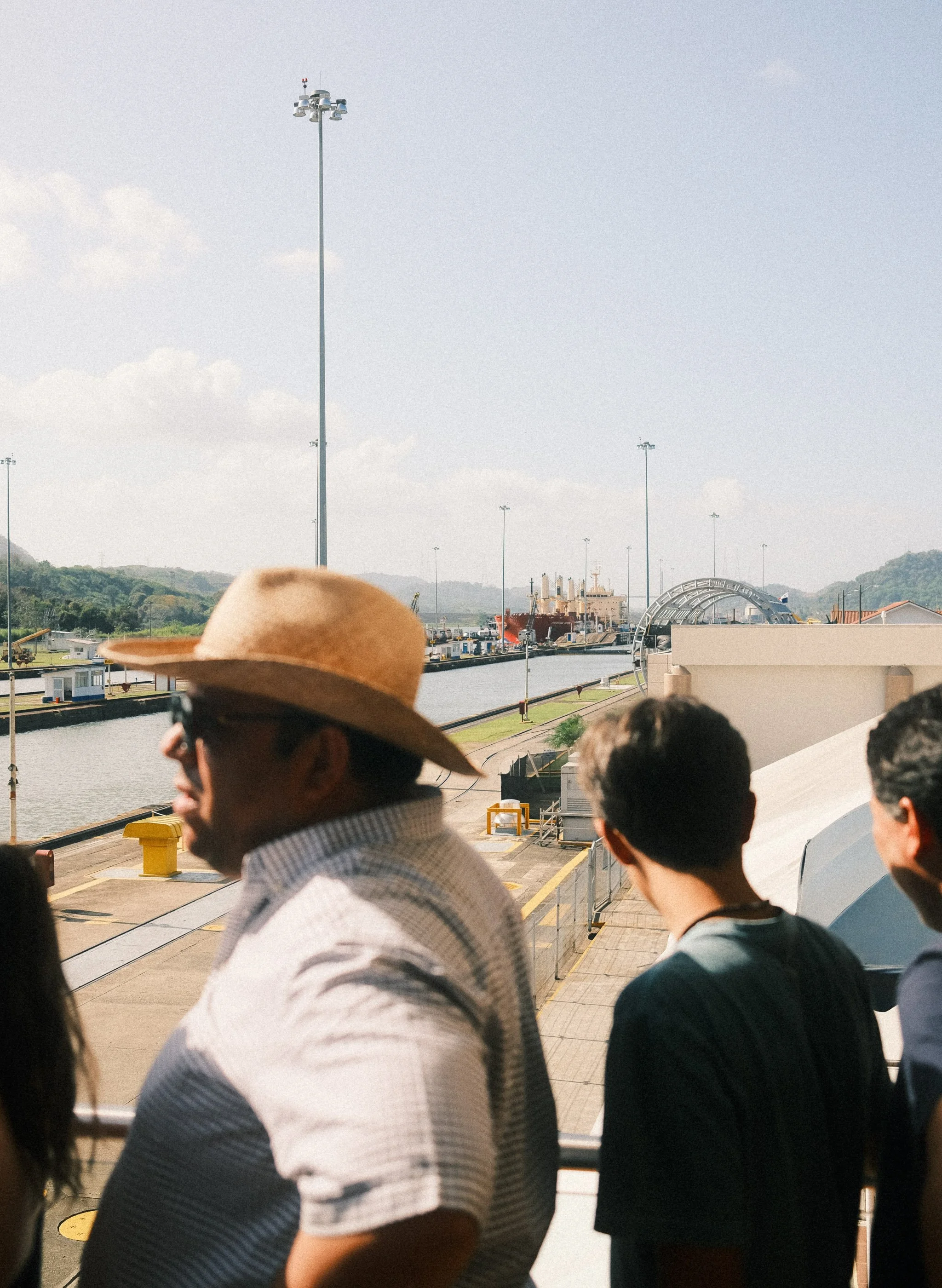 People waiting outdoors at a dock beside a waterway, with ships and industrial structures visible in the background under a partly cloudy sky.
