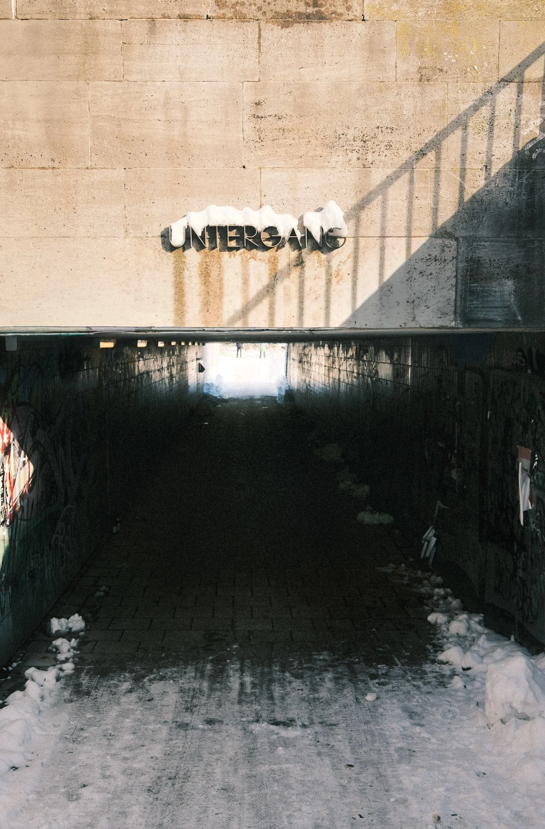 Entrance to a tunnel with the sign 'UNDERGANG' on a concrete wall, snow and graffiti on the sides, sunlight at the end of the tunnel.