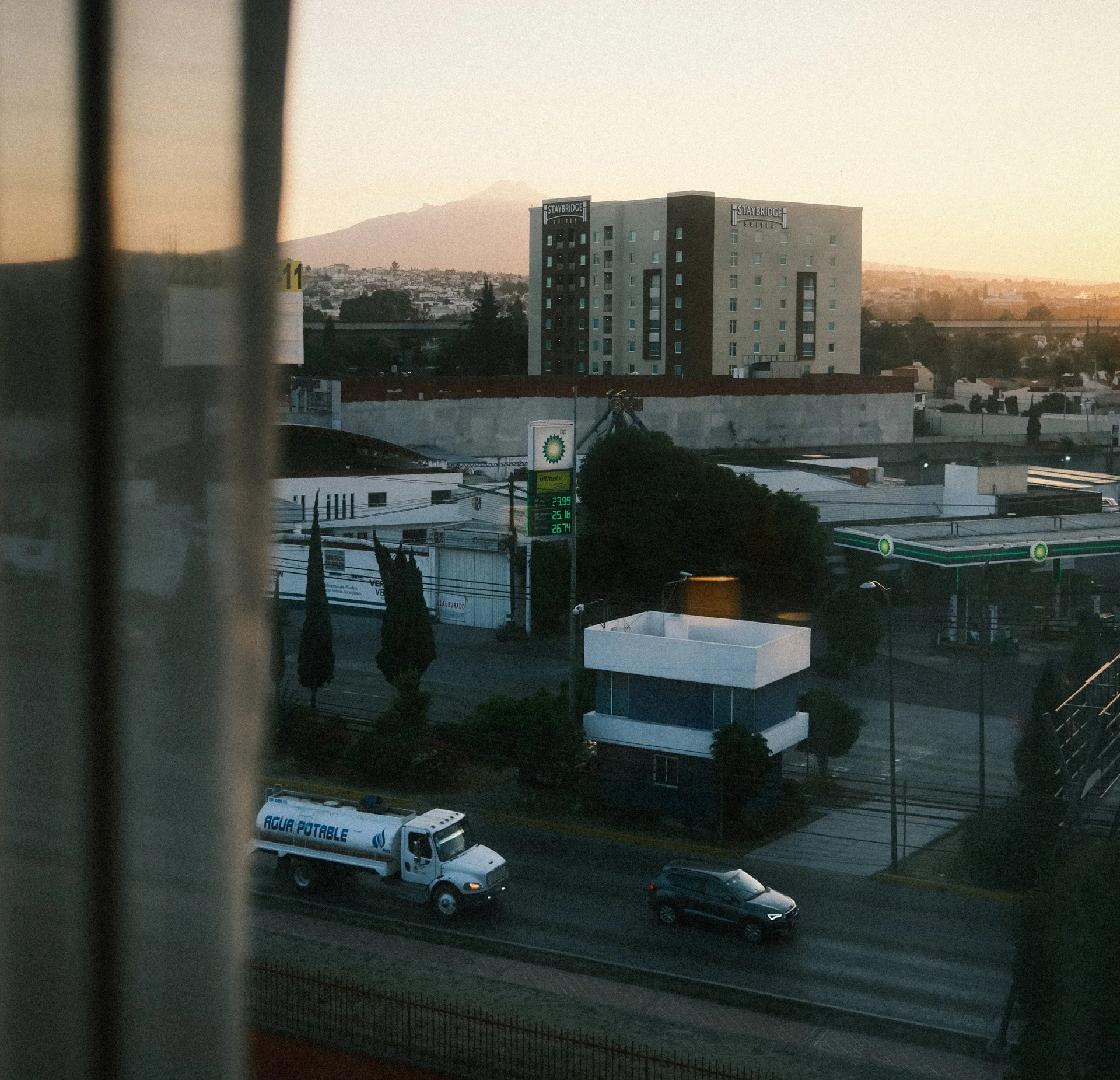 View of cityscape during sunset with buildings, a gas station, a water truck, and cars on the road, partially obscured by curtains.