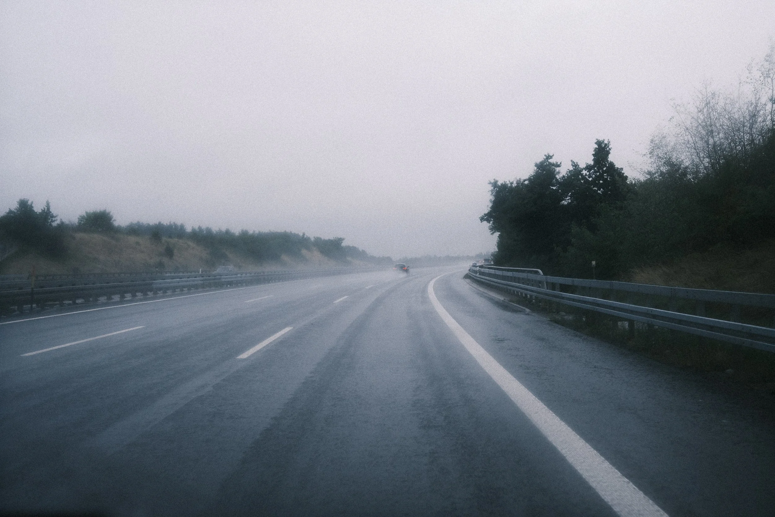 A wet, rainy highway with a car in the distance, surrounded by trees and guardrails on the side, under a cloudy, overcast sky.