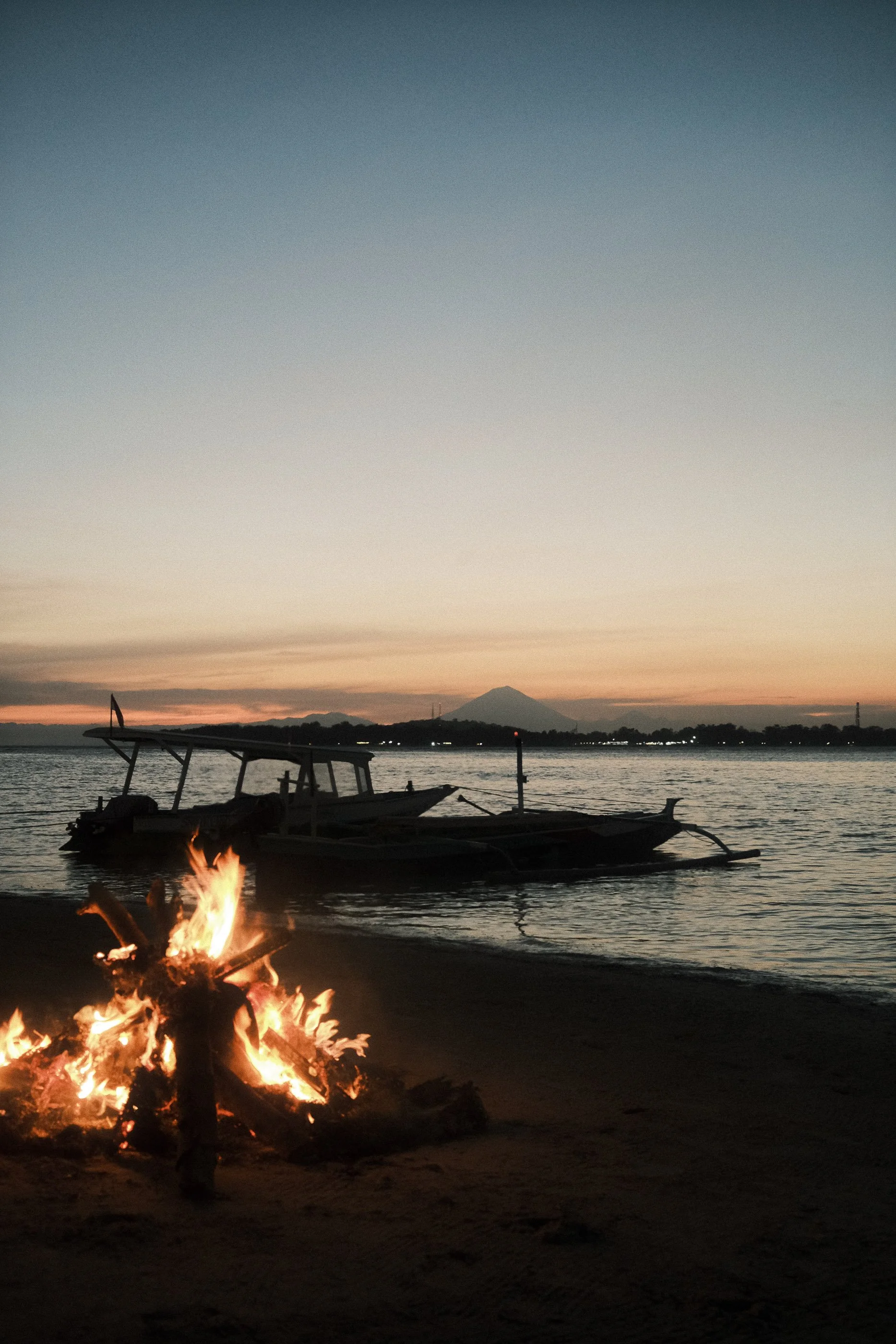 Sunset by the water with boats anchored near the shore, a campfire burning on the beach, and a mountain in the background.
