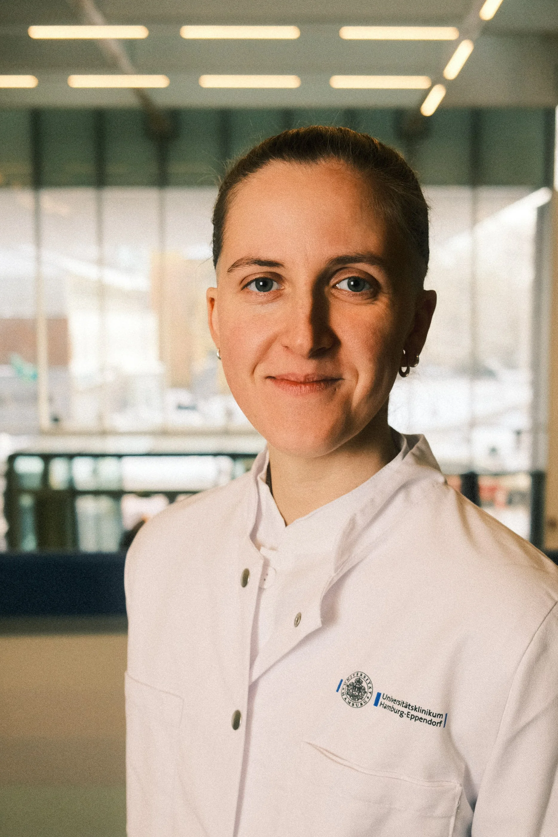 A person in a white medical coat standing indoors a hospital with large windows in the background.