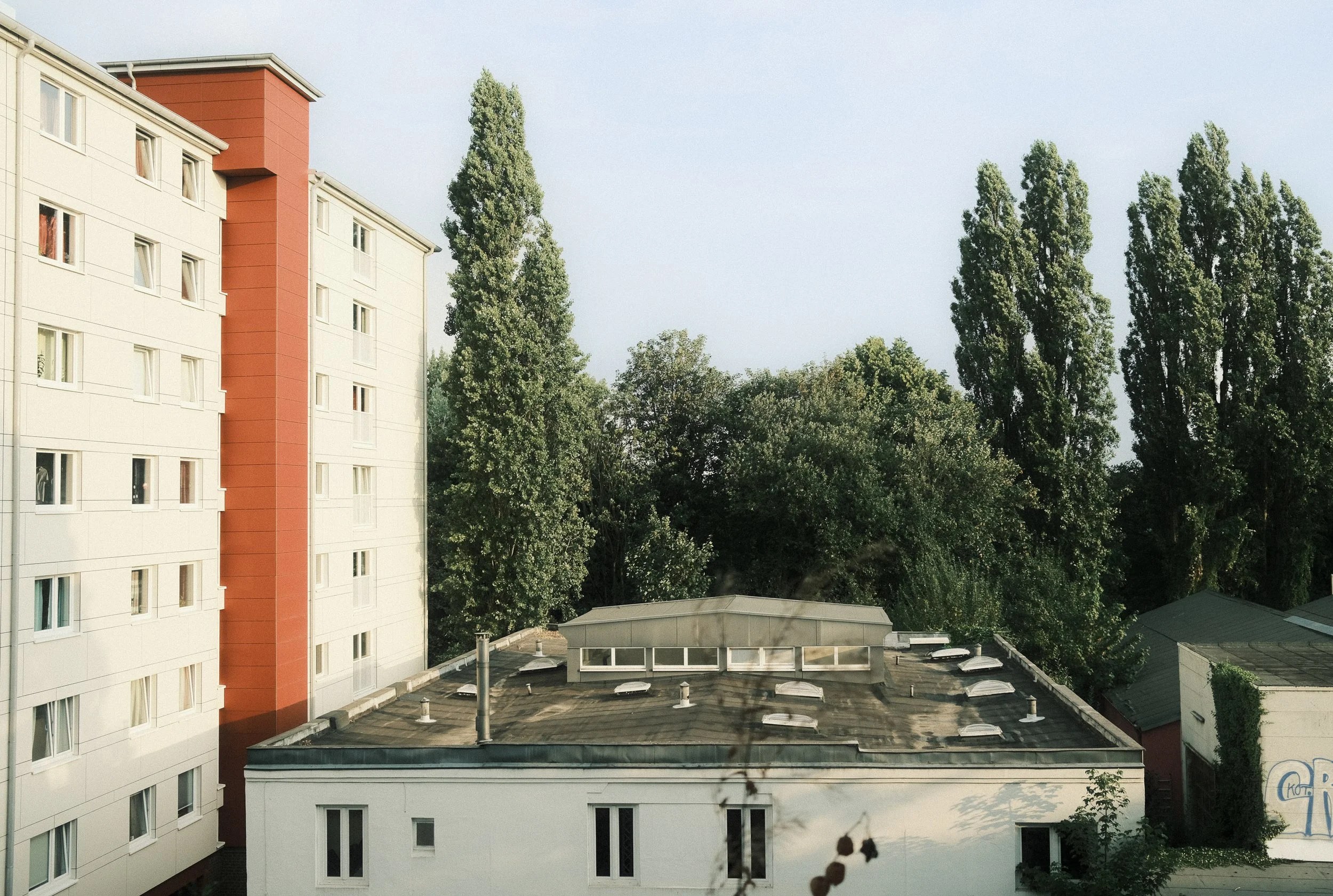 Apartment building with multiple windows, a red vertical section, and surrounded by tall green trees and a flat roof with skylights.