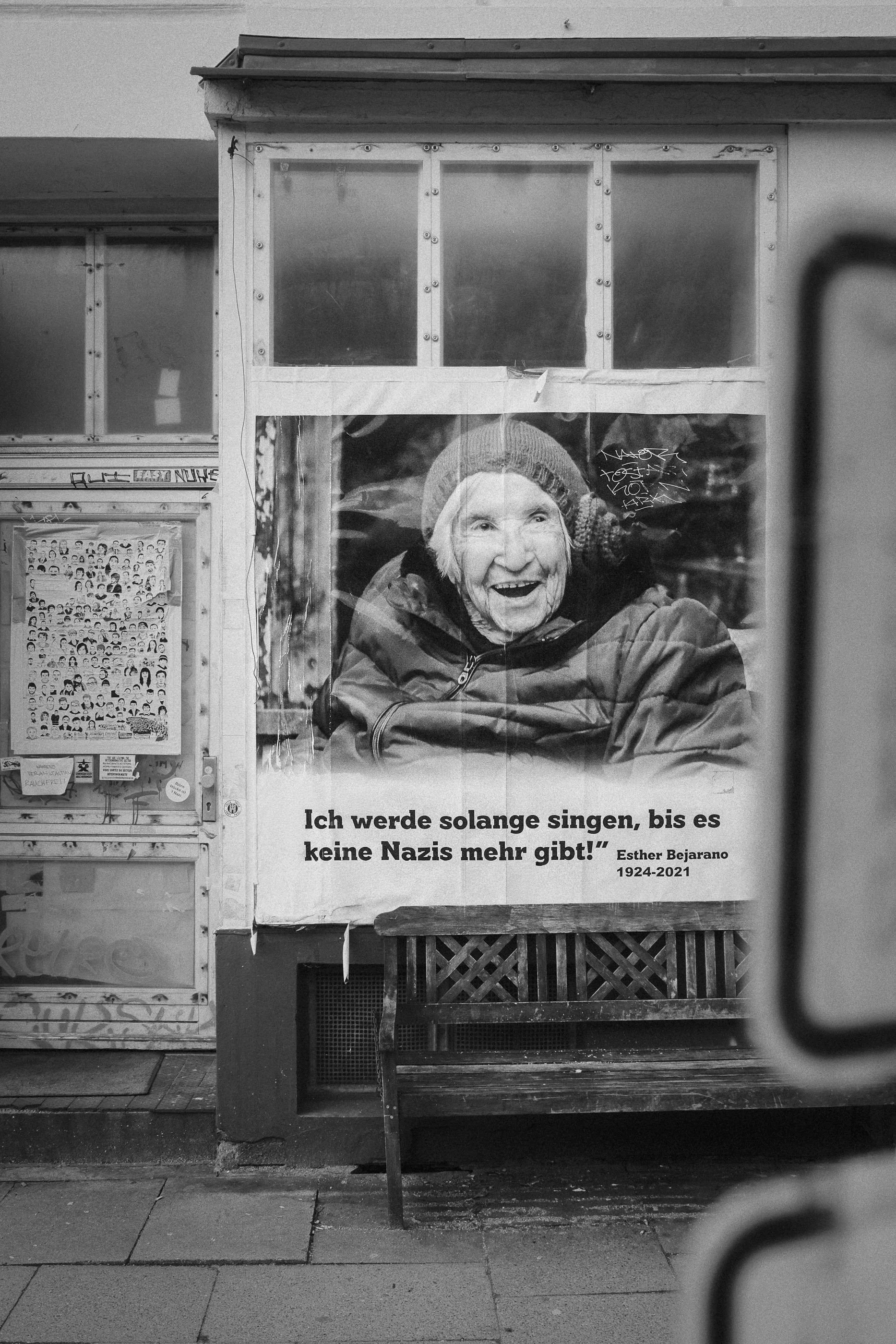 A black and white photo of a poster featuring an elderly woman smiling, wearing a beanie and winter clothing. The poster includes a quote in German: 'Ich werde solange singen, bis es keine Nazis mehr gibt!' attributed to Esther Bejarano (1924-2021). 