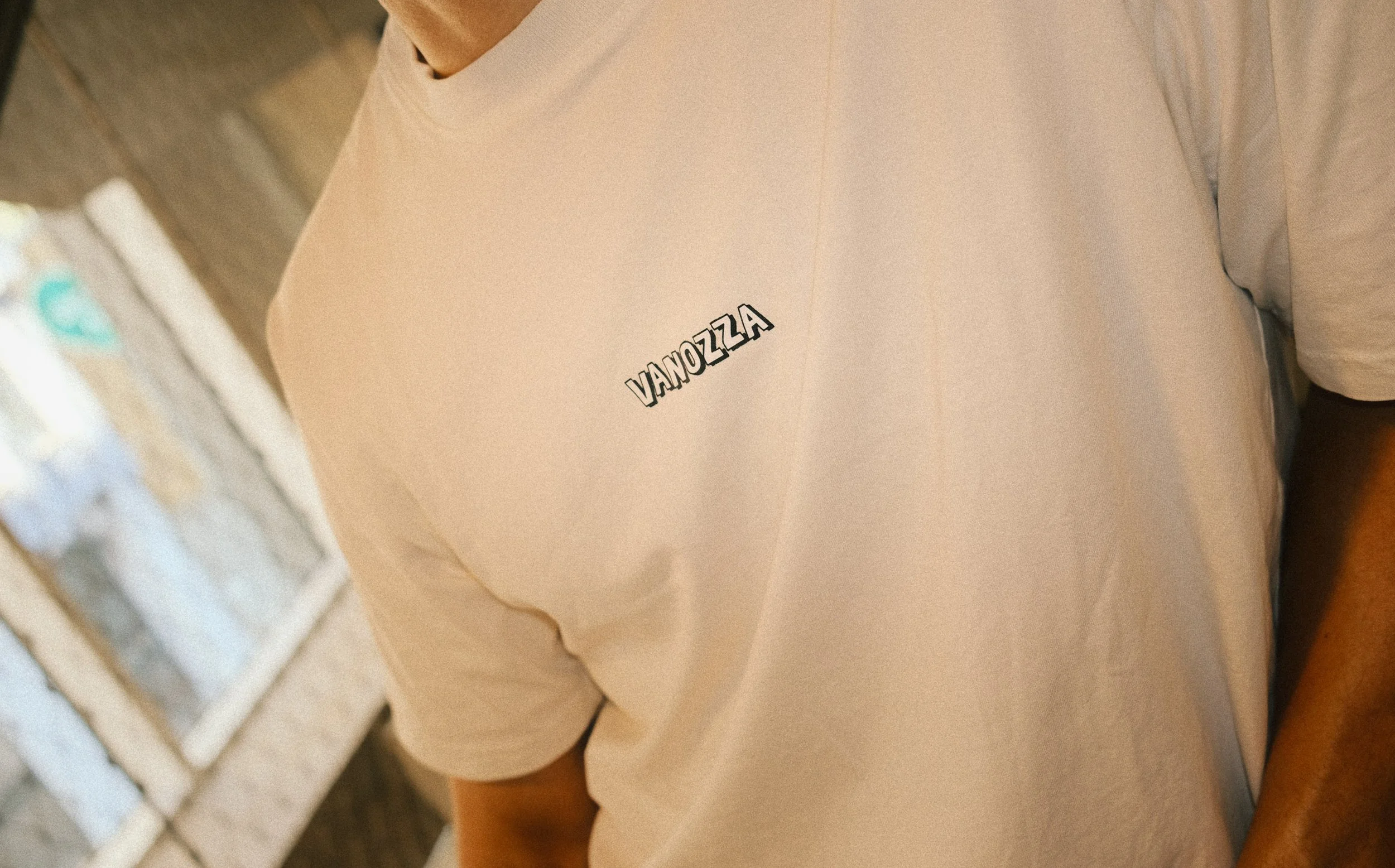 Close-up of a person wearing a beige T-shirt with the word "VANOZZA" written on it, standing indoors near a window.