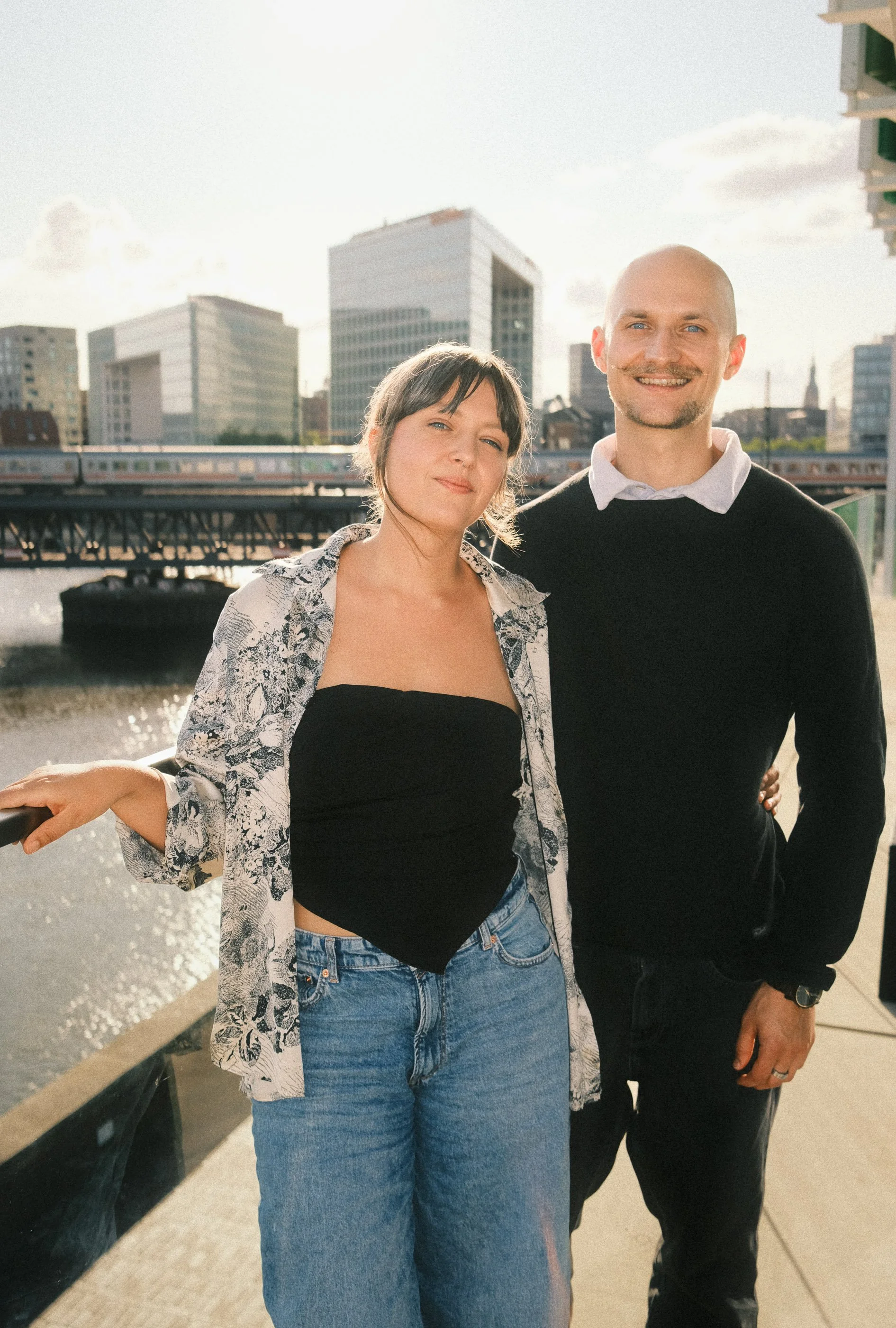 A woman and a man standing together outdoors near a body of water with city buildings in the background, smiling at the camera.
