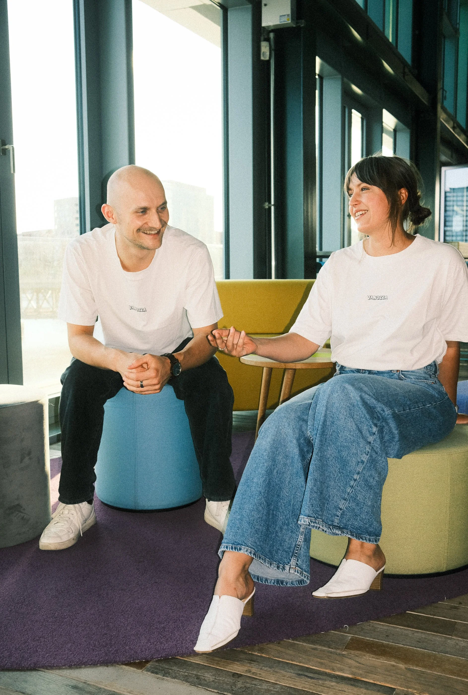 A man and woman sitting in a modern office lounge, smiling and holding hands, during a casual conversation.