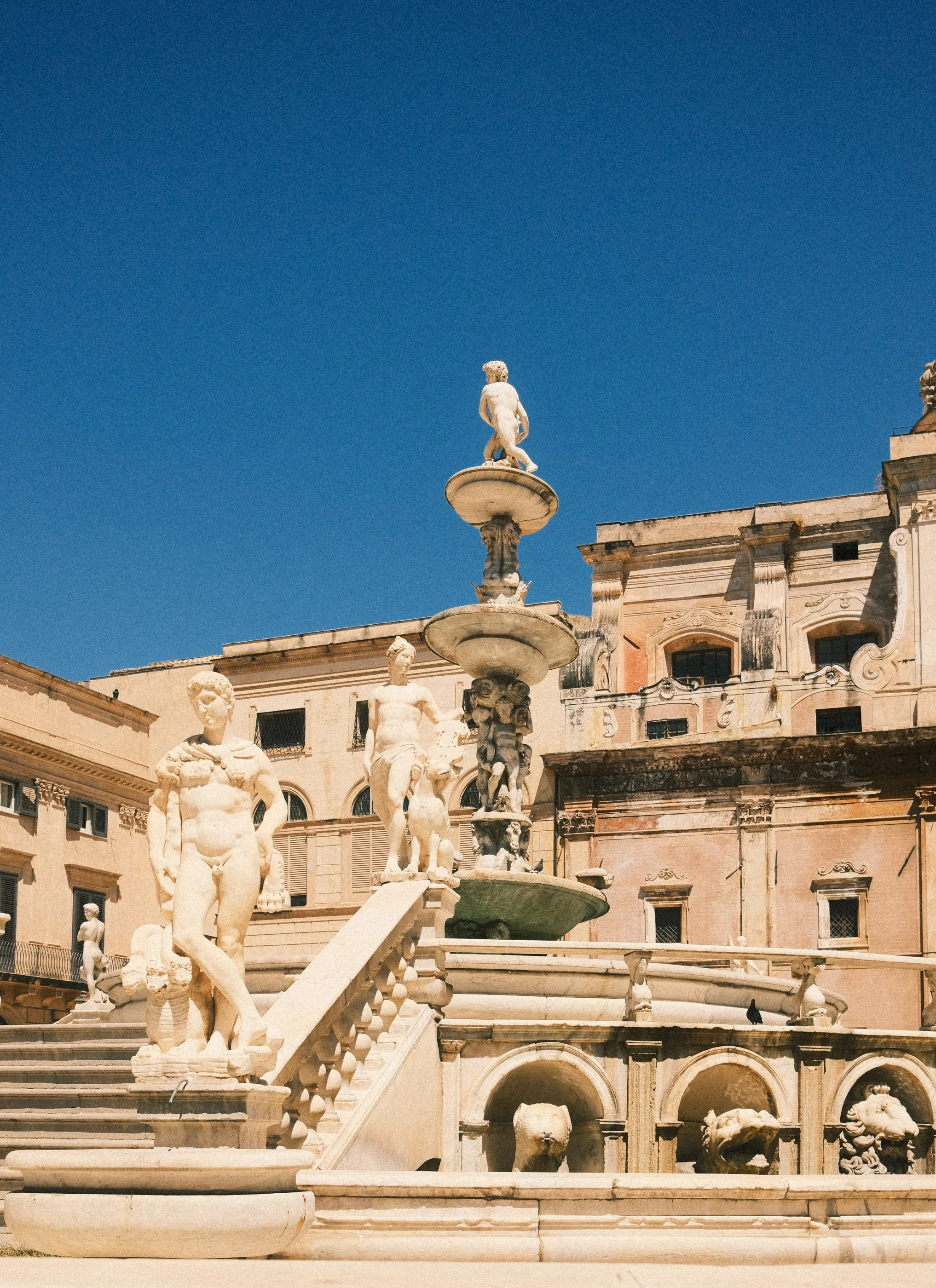 An ornate marble fountain with multiple sculpted figures and statues, including children and animals, set against historic buildings under a clear blue sky.