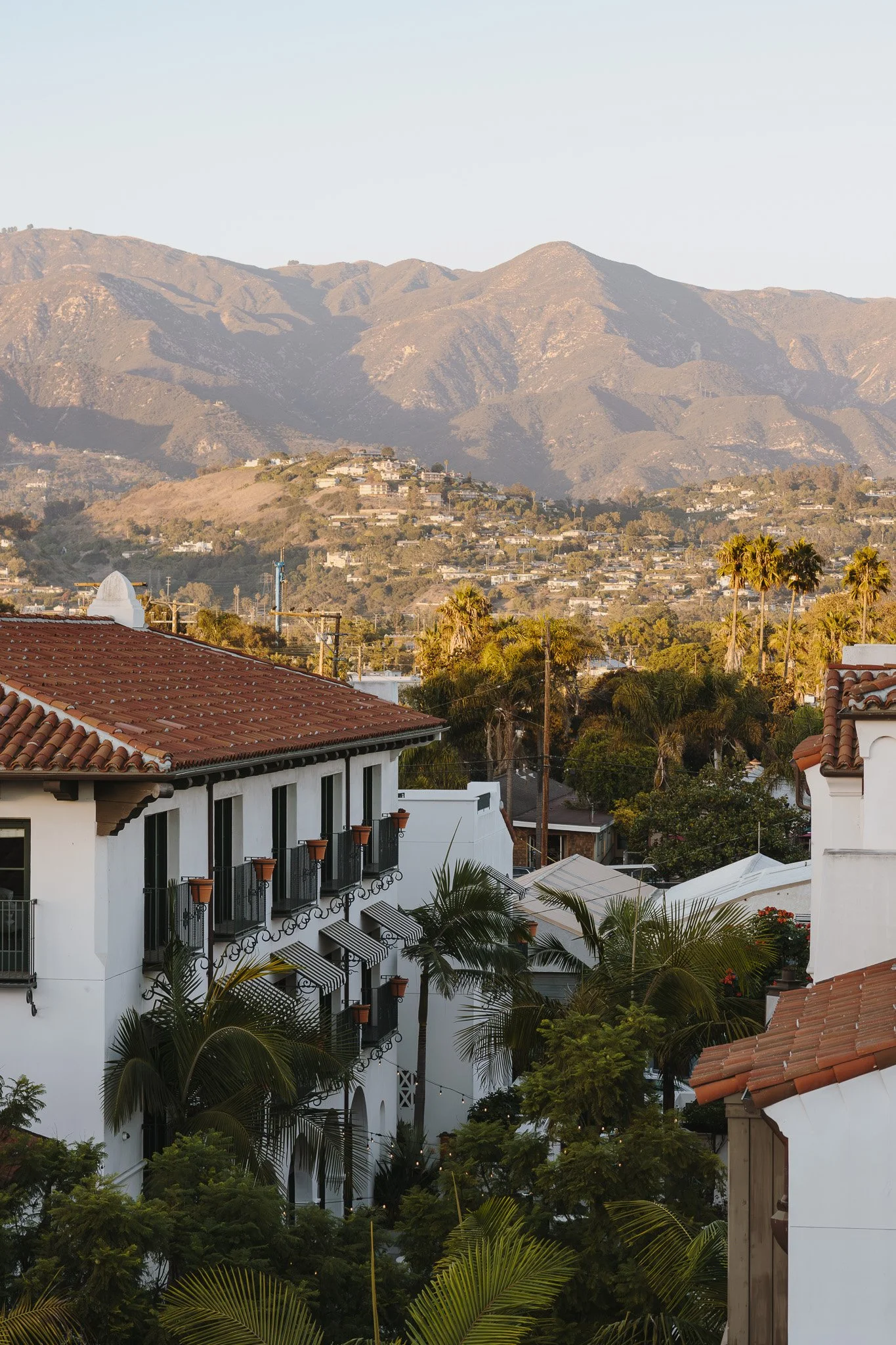 A scenic view of a hillside residential area with white buildings featuring red-tiled roofs, surrounded by lush green trees and tall palm trees, with mountains in the background under a clear sky.