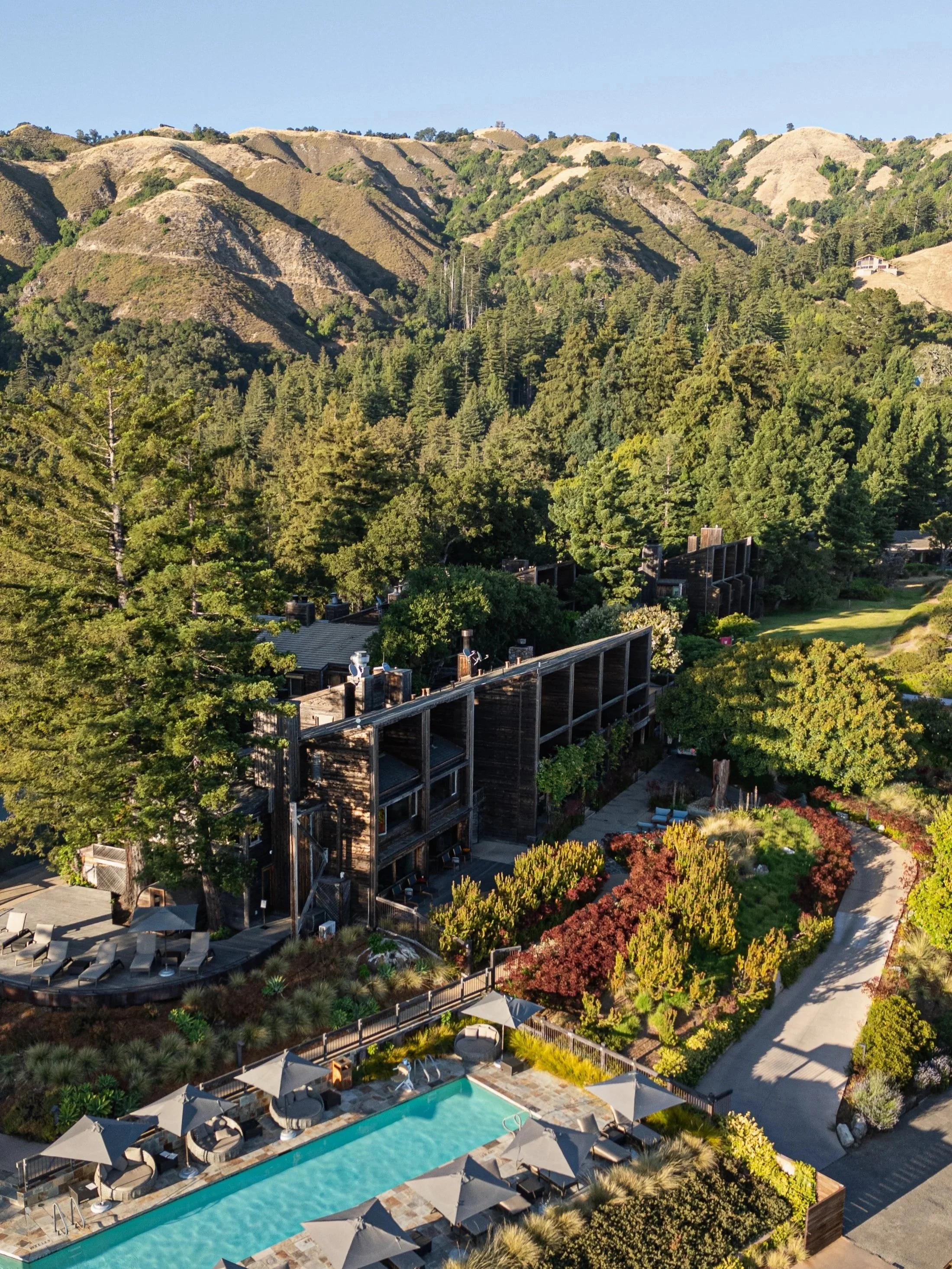 Aerial view of a hiking trail, a swimming pool with lounge chairs and umbrellas, surrounded by lush greenery and trees, with mountain hills in the background.