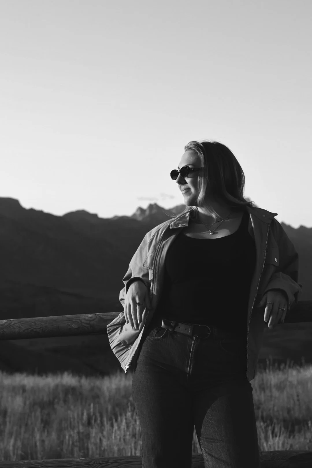 A woman standing outdoors near a wooden fence, wearing sunglasses and a jacket, with mountains and a clear sky in the background.