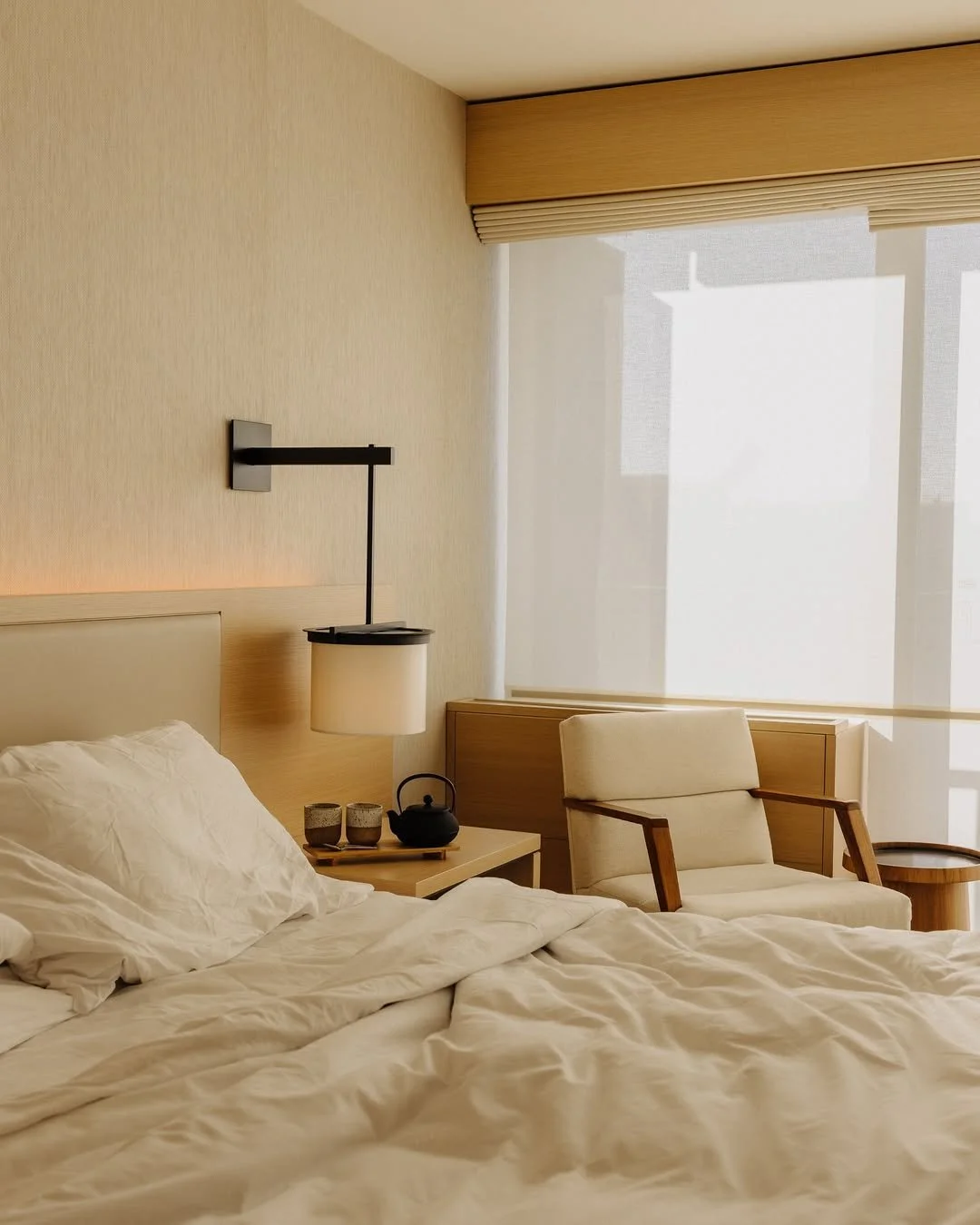Hotel room with a bed, nightstand, armchair, side table, and large window with rolled shades.