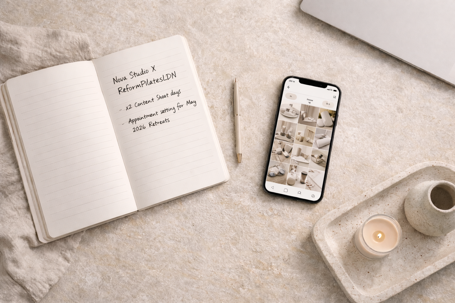 Open notebook with handwritten notes, a decorative tray with a lit candle and a small ceramic vase, a smartphone displaying a grid of neutral-toned home decor items, and a closed laptop on a textured surface.