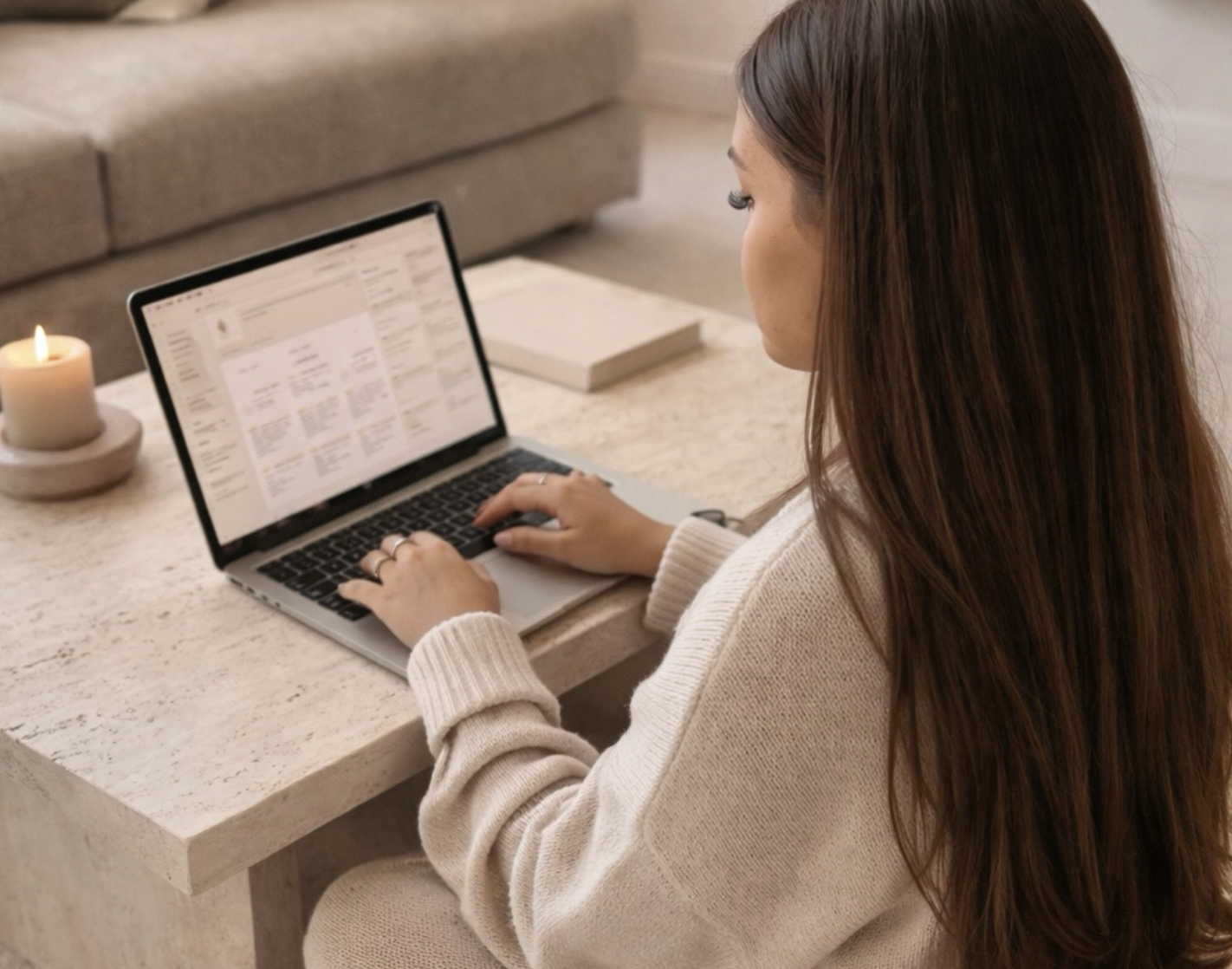 Woman with long brown hair working on a laptop at a light-colored table, with a candle and a book nearby.