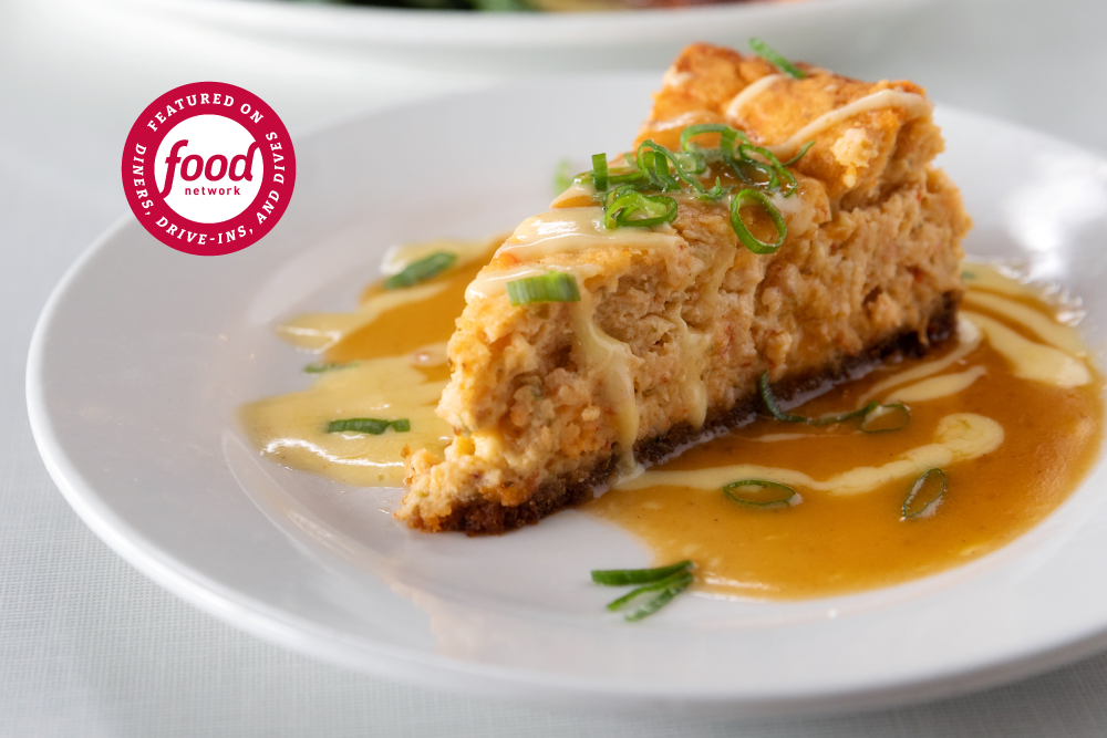 Slice of savory, cheesy Crawfish Cheesecake topped with green onions, served with a creamy gravy and drizzled with hollandaise on a white plate.
