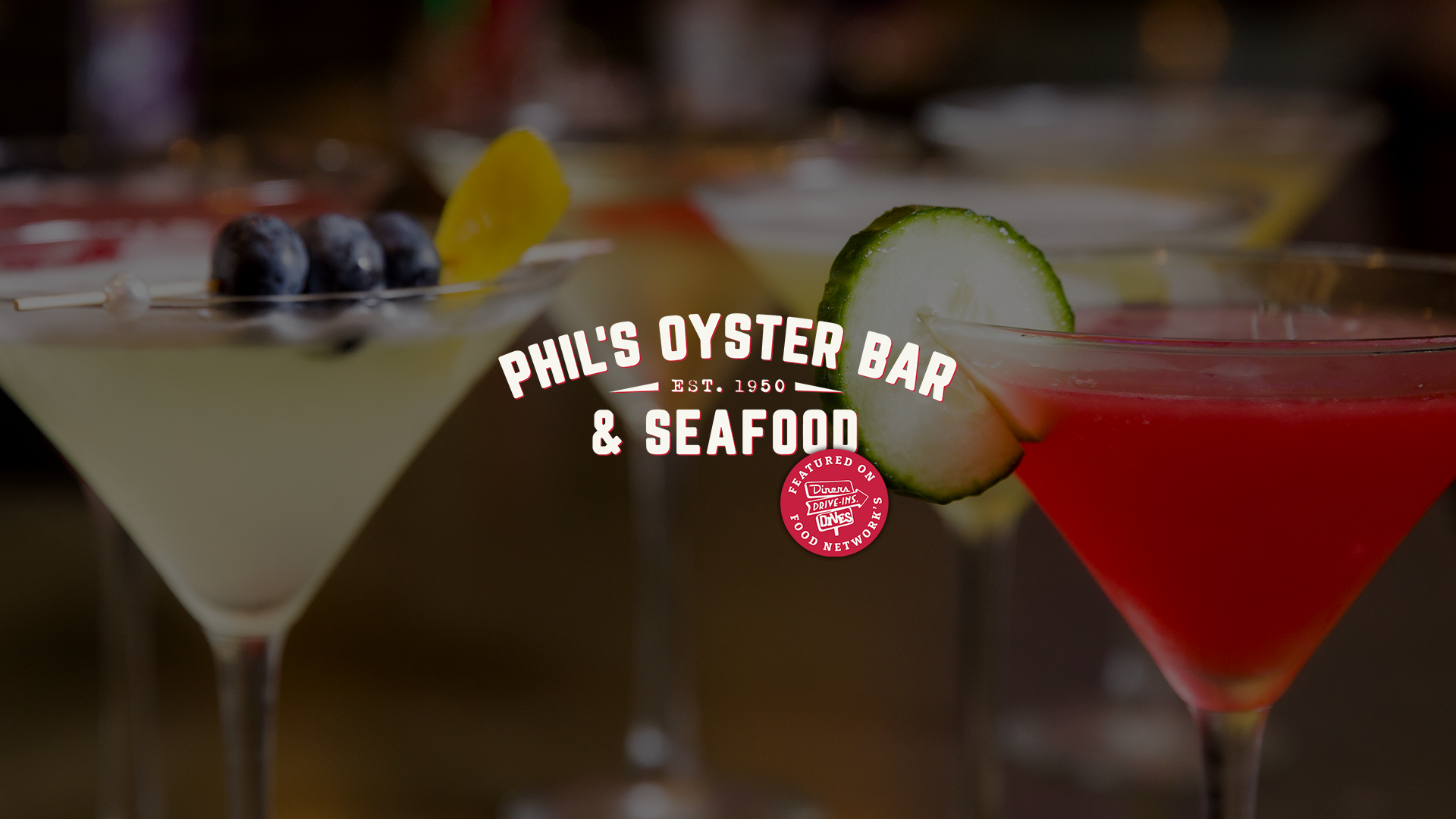 Selection of colorful specialty martinis and cocktails at the Phil's Oyster Bar & Seafood bar. Overlaid text reads 'Phil's Oyster Bar & Seafood, Est. 1950.' with Food Network's Diners, Drive-Ins, Drives logo.