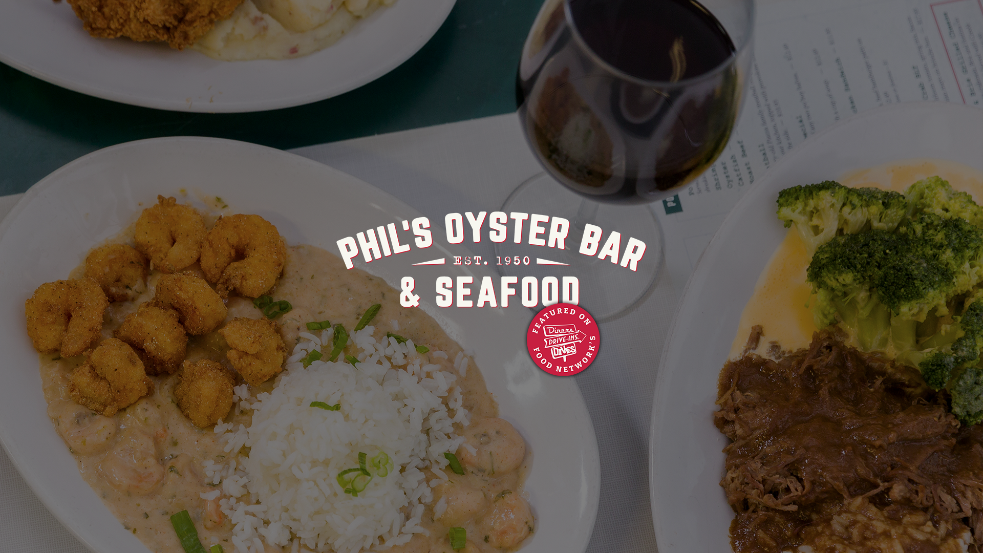 Table spread with plates featuring fried shrimp, roast beef with broccoli, and a glass of red wine. Overlaid text reads 'Phil's Oyster Bar & Seafood, Est. 1950.' with Food Network's Diners, Drive-Ins, Drives logo.