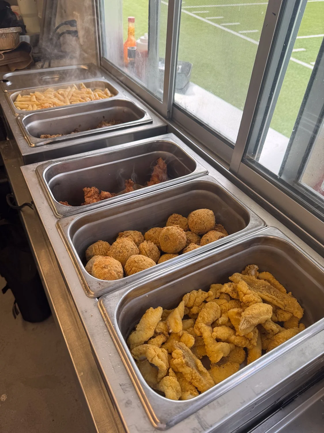 Buffet-style tray with fried chicken, fried fish, boudin balls, pasta, and cooked meat inside a food truck, near a window overlooking a sports field.