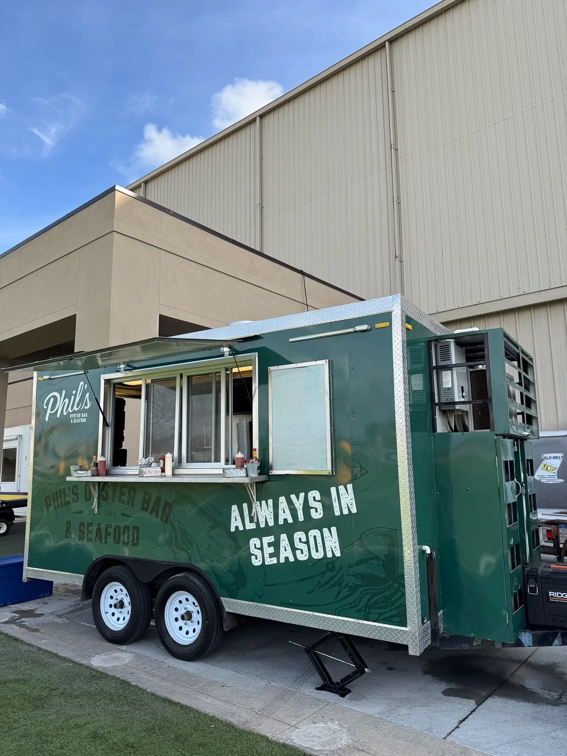 Green food truck named Phil's Oyster Bar & Seafood with 'Always in Season' on the side, parked near a beige building on a paved area, under a blue sky with some clouds.