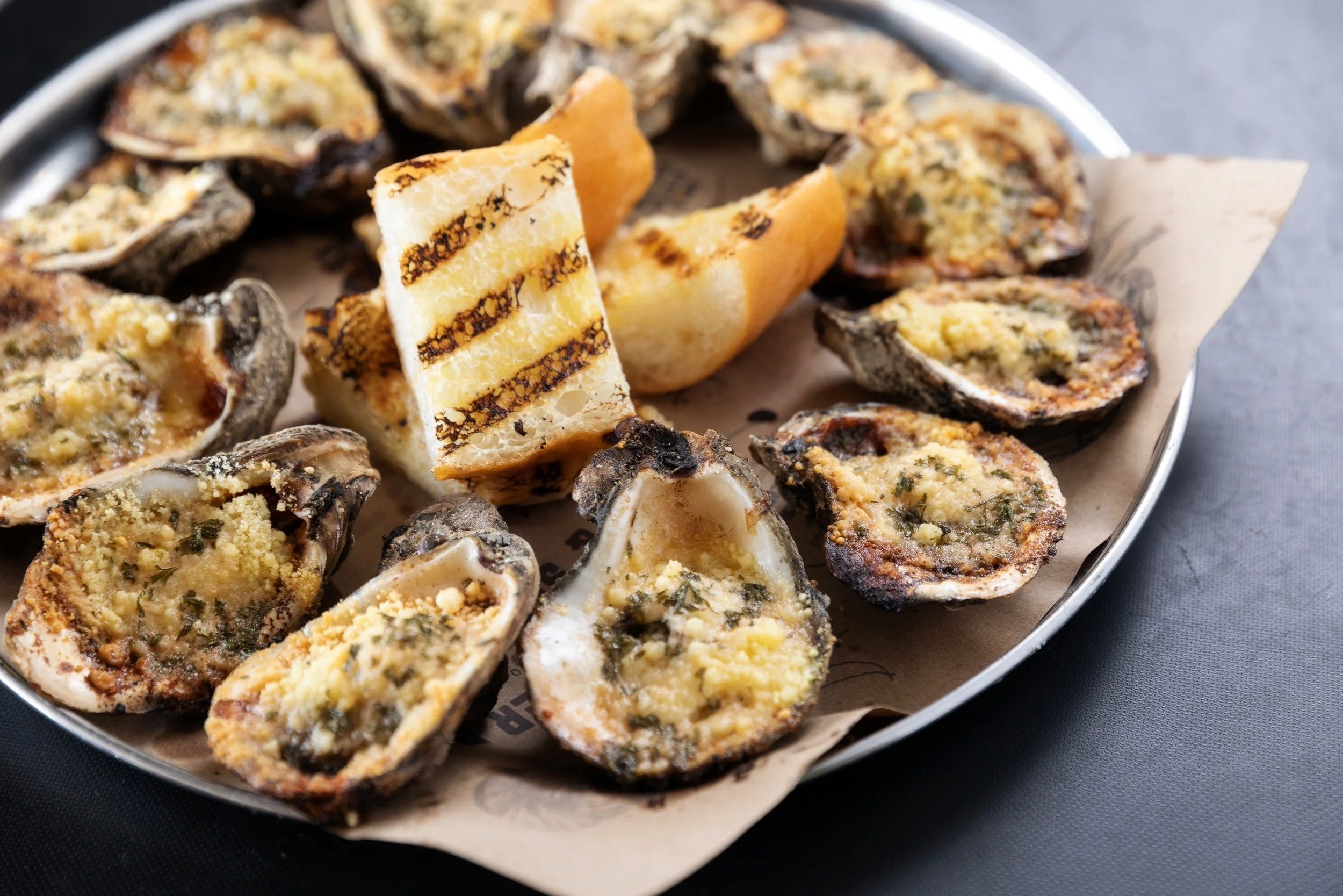 A metal tray with chargrilled oysters topped with cheese and butter, with a slice of French bread on top, placed on a gray surface.