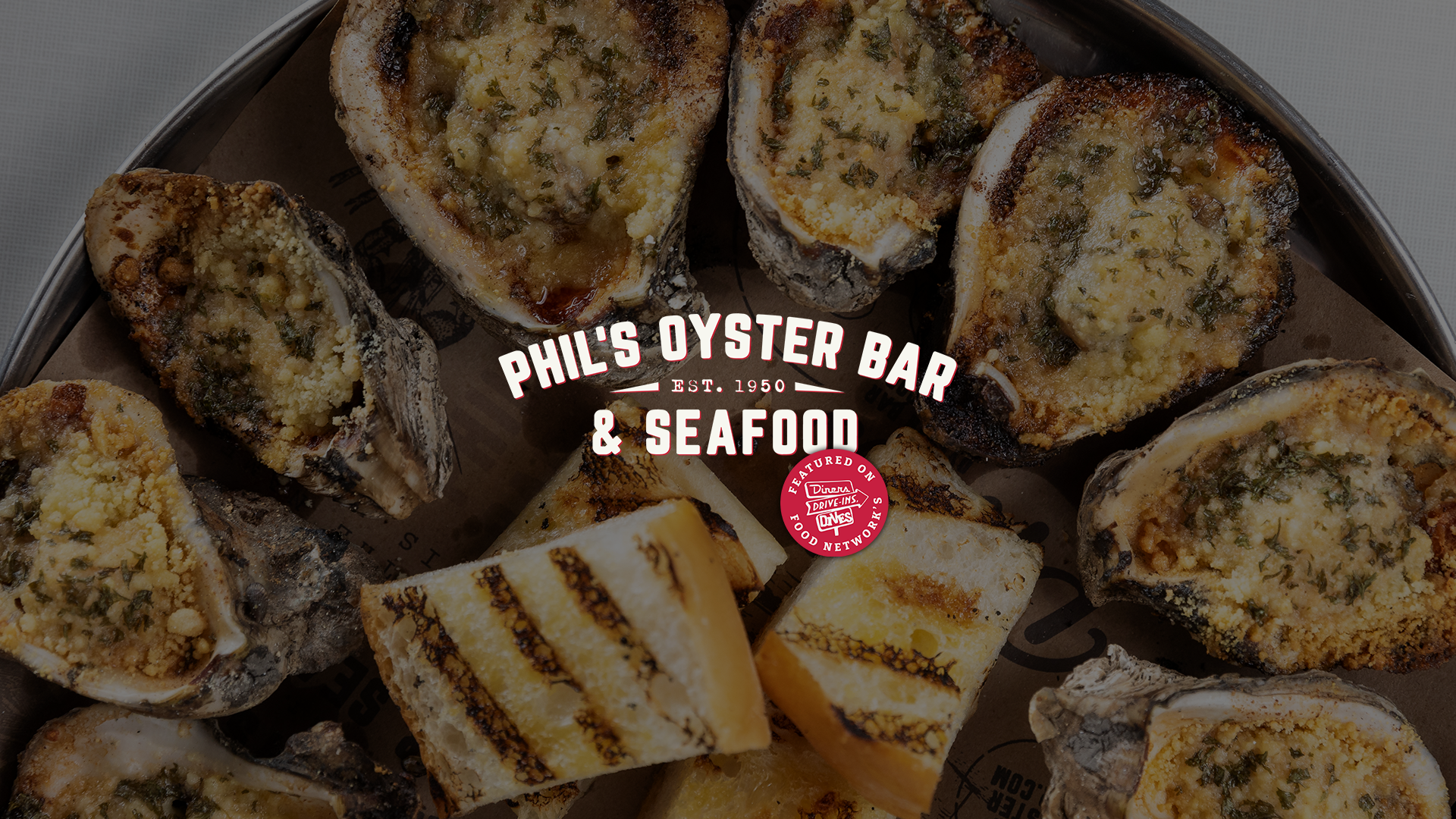 Close-up of chargrilled oysters topped with cheese and butter. Overlaid text reads 'Phil's Oyster Bar & Seafood, Est. 1950.' with Food Network's Diners, Drive-Ins, Drives logo.