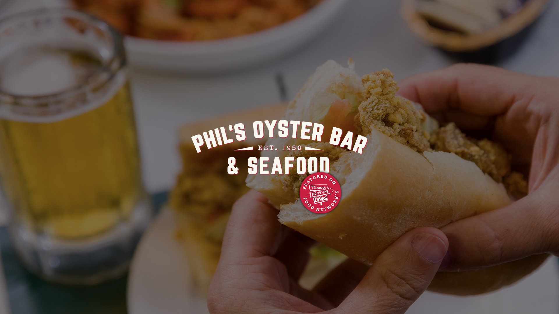 Close-up of a fried shrimp po-boy, held by a person. In the background, a glass of beer and a plate of food are visible. Overlaid text reads 'Phil's Oyster Bar & Seafood, Est. 1950.' with Food Network's Diners, Drive-Ins, Drives logo.