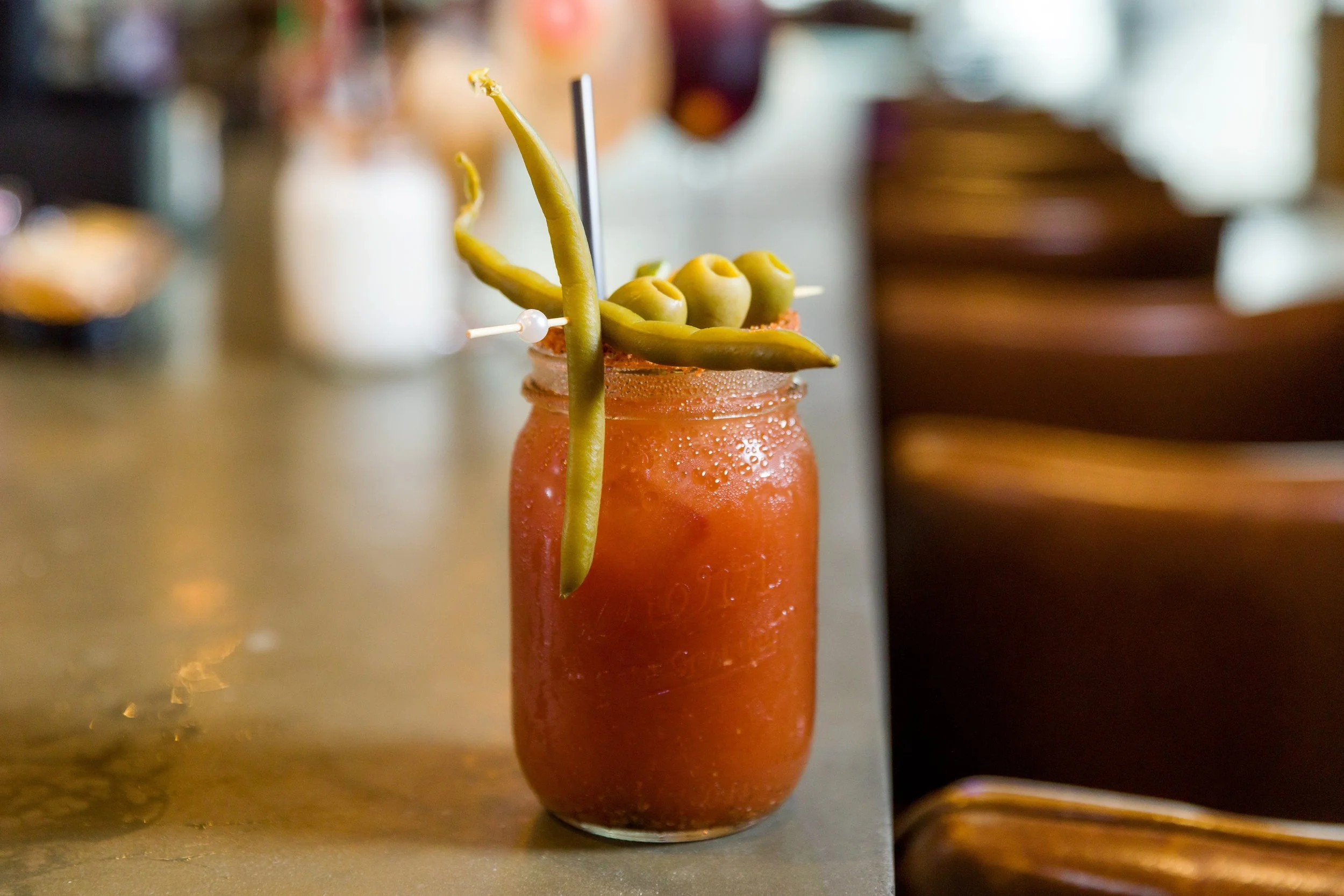 Bloody Mary cocktail garnished with green beans, olives, and a straw on a bar counter.
