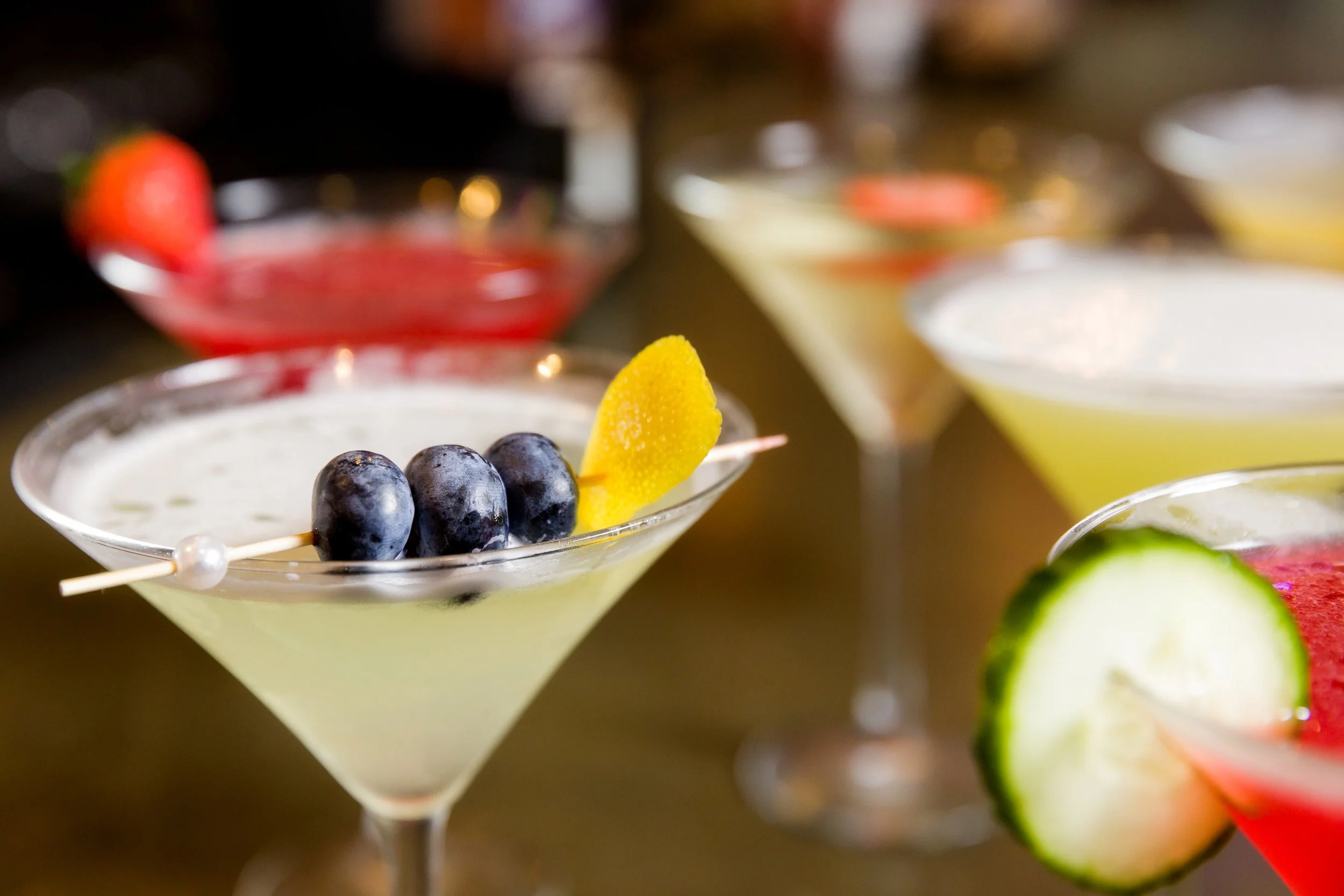 Assorted colorful cocktails garnished with fruit, including blueberries, lemon, cucumber, and strawberry.