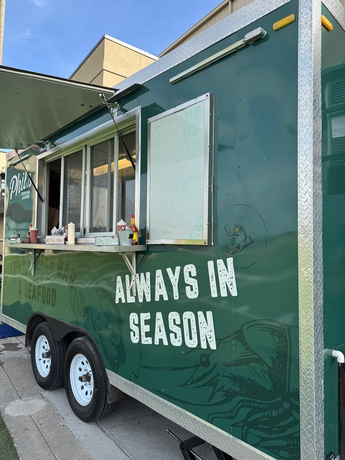 Green food truck named Phil's Oyster Bar & Seafood with 'Always in Season' on the side, parked near a beige building on a paved area, under a blue sky with some clouds.
